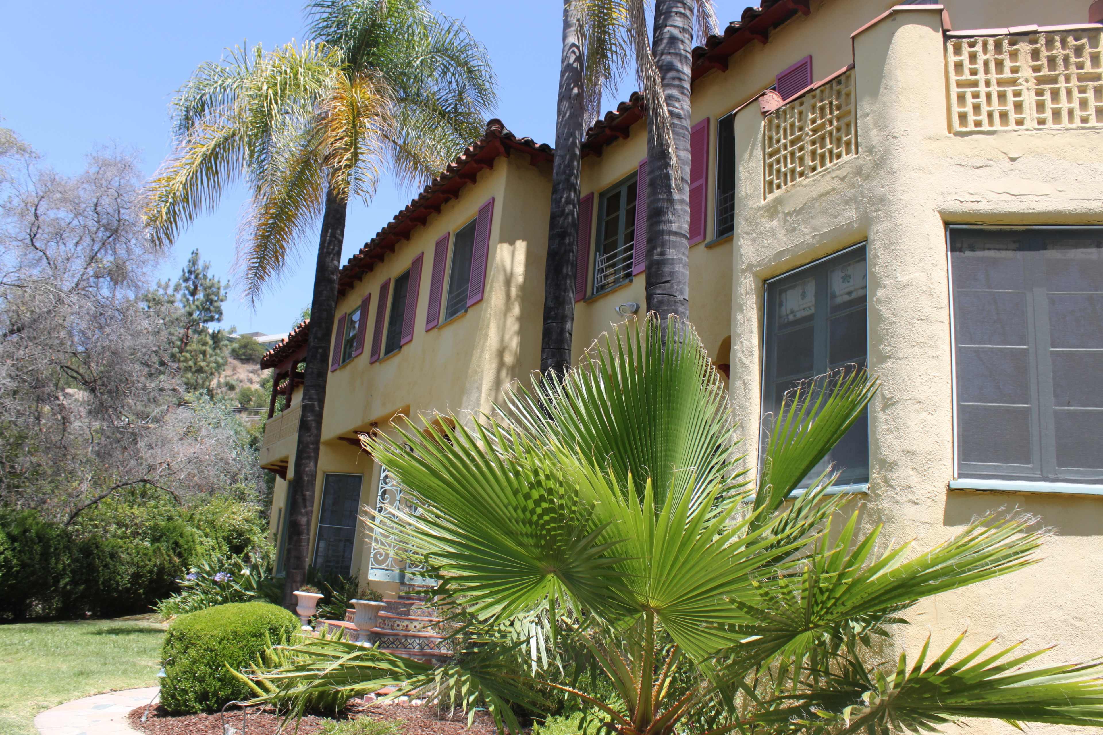 The image shows a two-story yellow house with red trim, surrounded by palm trees and green shrubbery.
