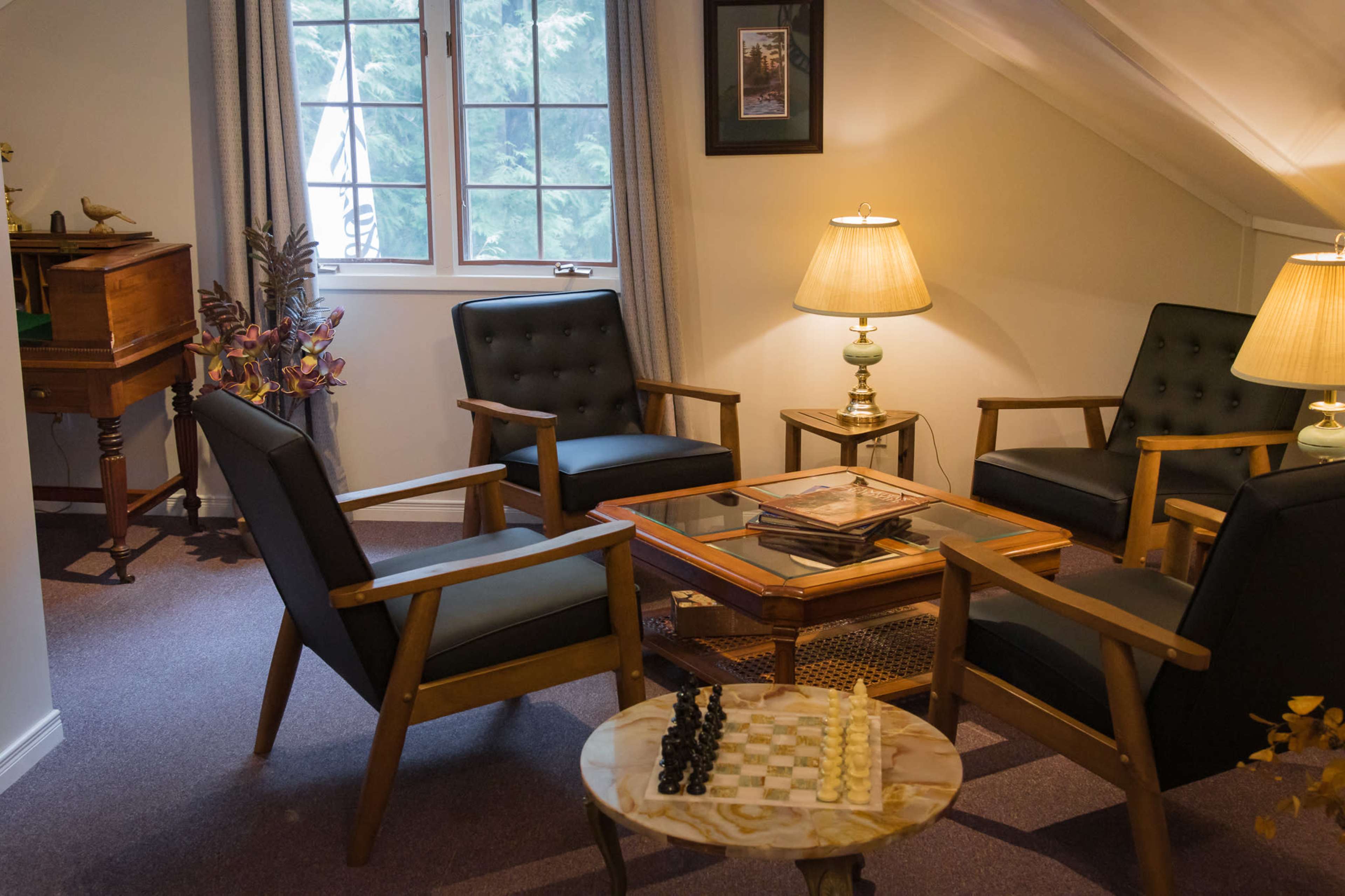 A cozy sitting area with two armchairs and a coffee table, featuring a chess set, alongside a piano in a well-lit room.