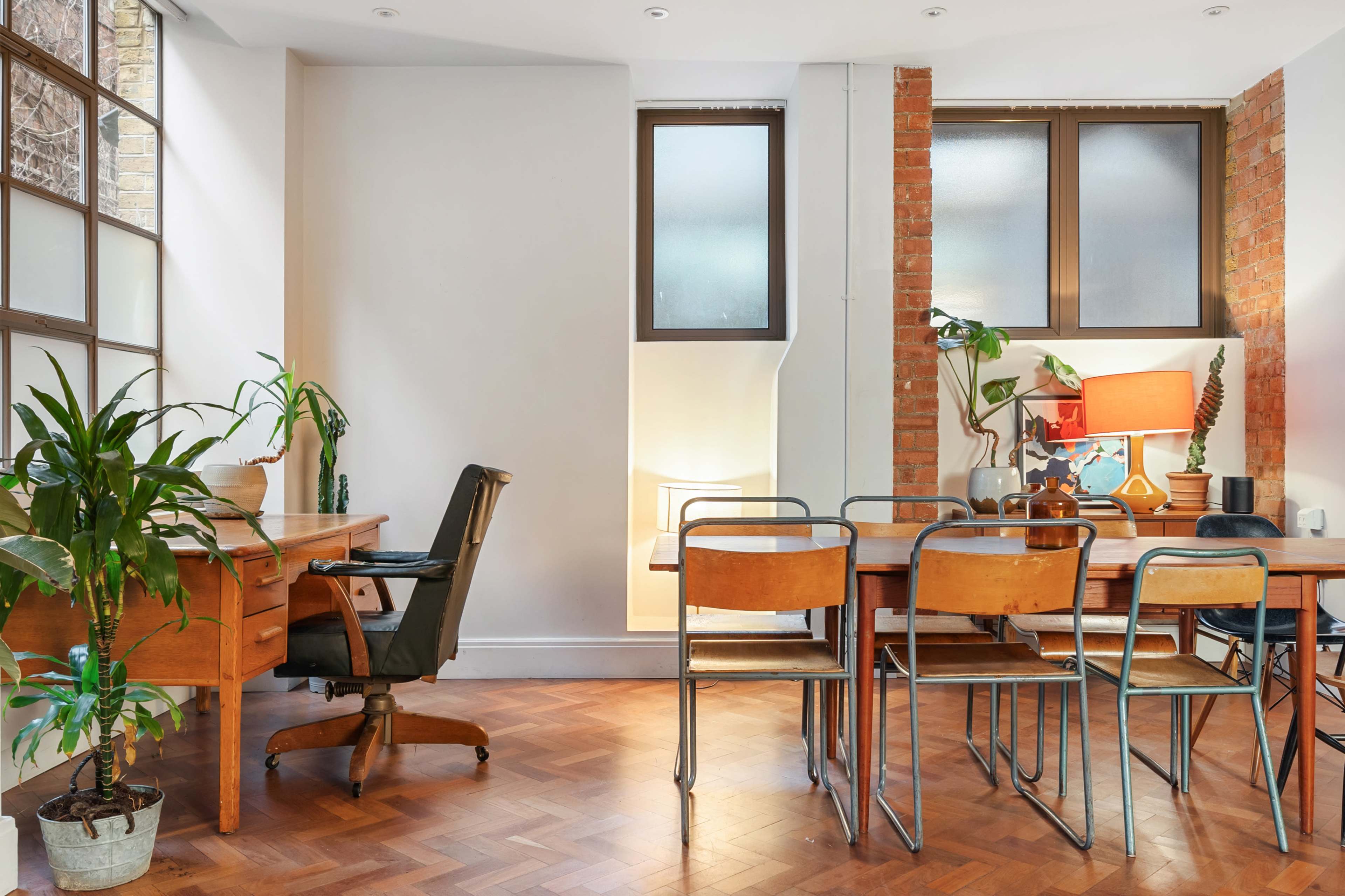 The image shows a bright, spacious room with a wooden desk and chair on one side and a large dining table with metal chairs on the other, featuring plants and decorative items.