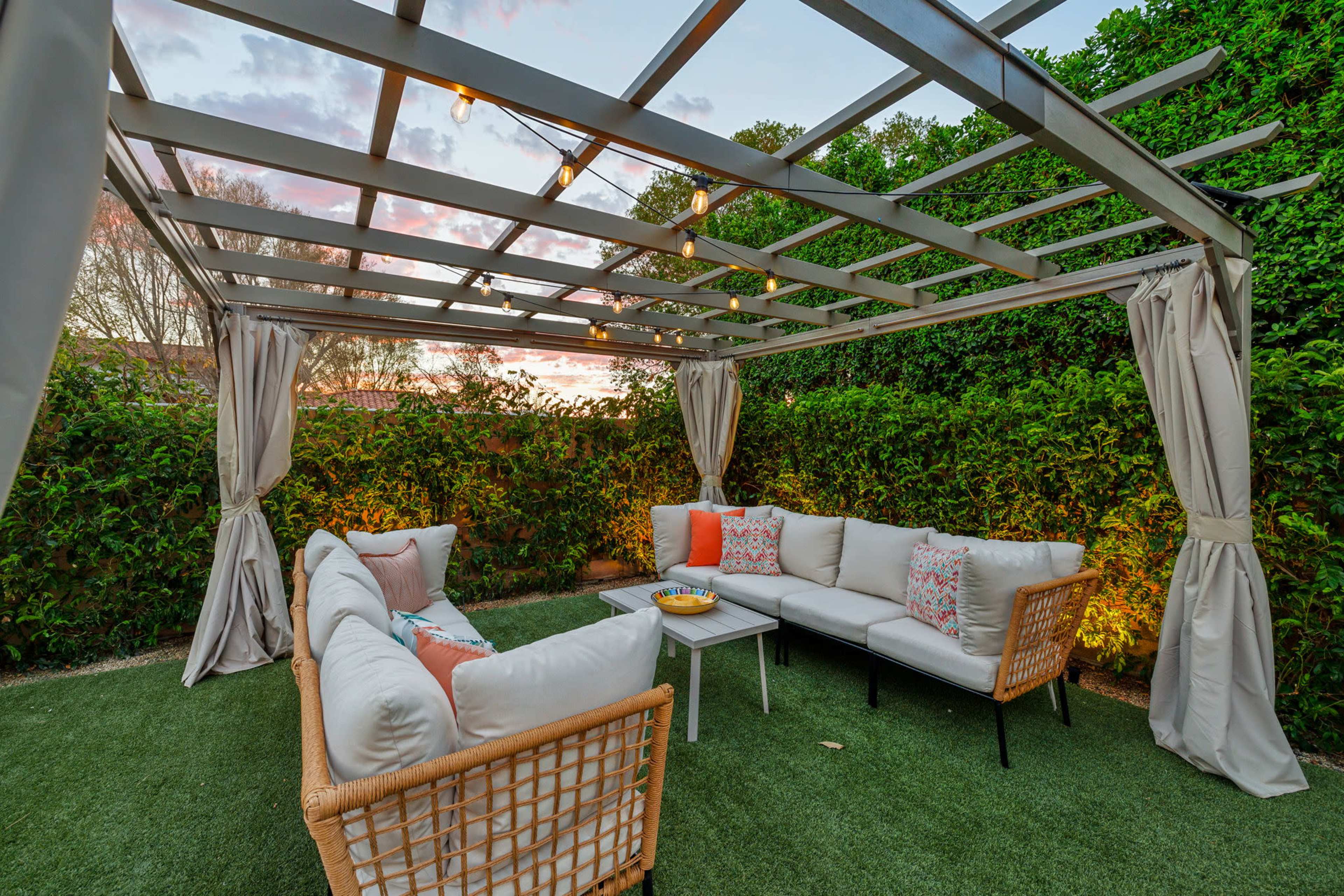 A cozy outdoor seating area is framed by a pergola with curtains, featuring a sectional sofa with decorative pillows and a small table.