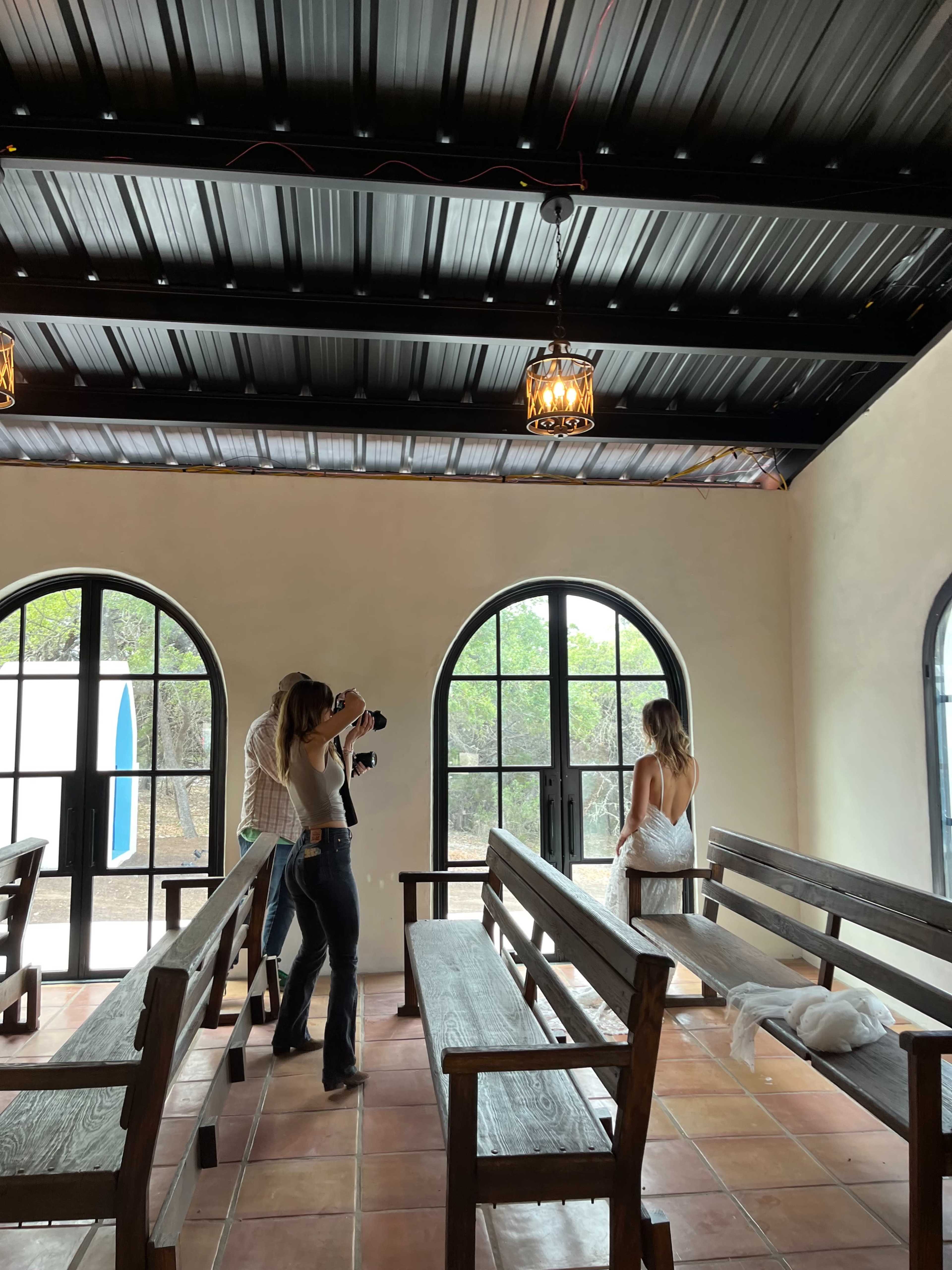 Two photographers are capturing images of a bride in a wedding dress inside a chapel with arched windows and wooden benches.