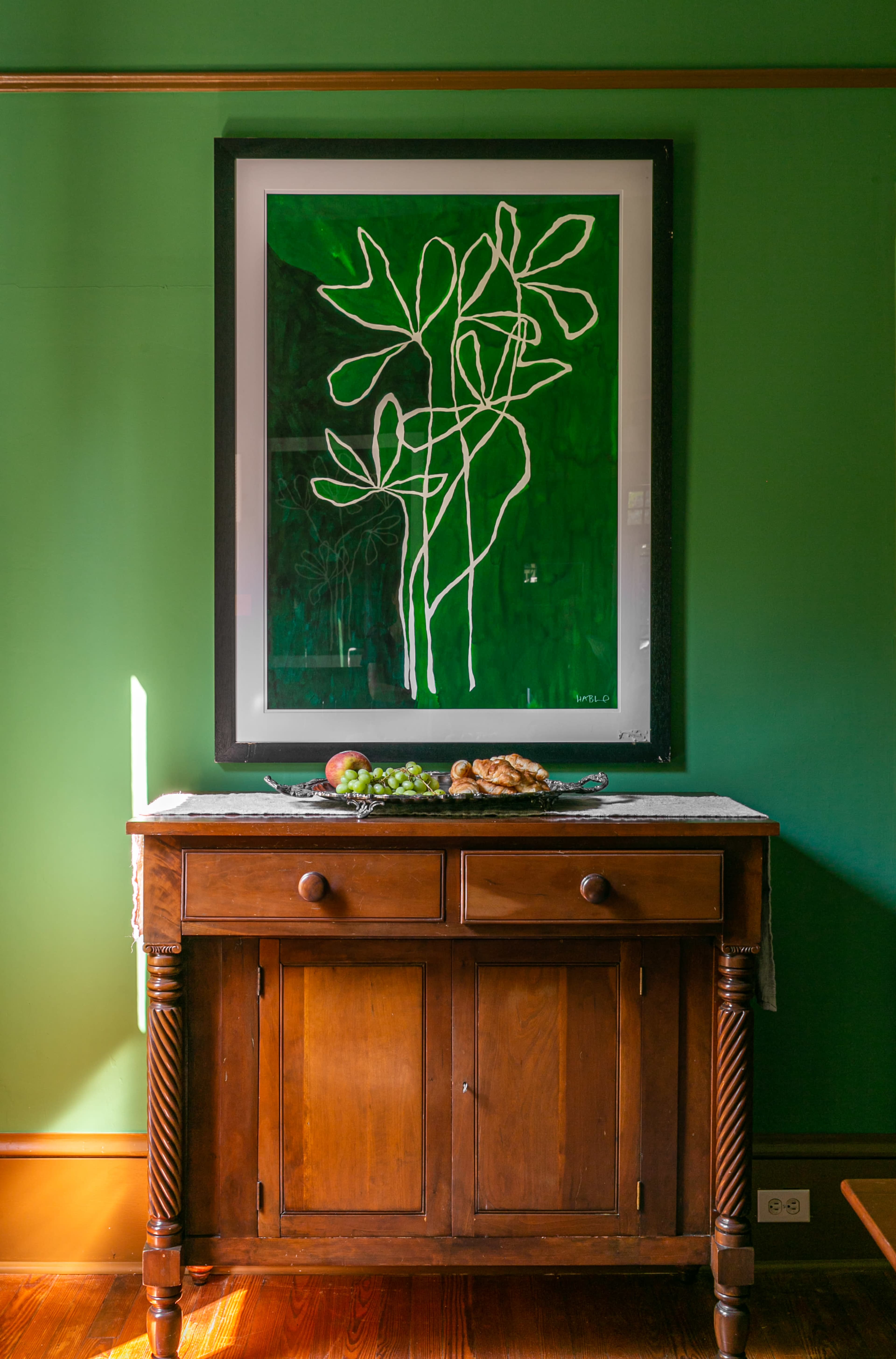 A wooden sideboard with a tablecloth displays a bowl of fruit and pastries beneath a large framed artwork on a green wall.