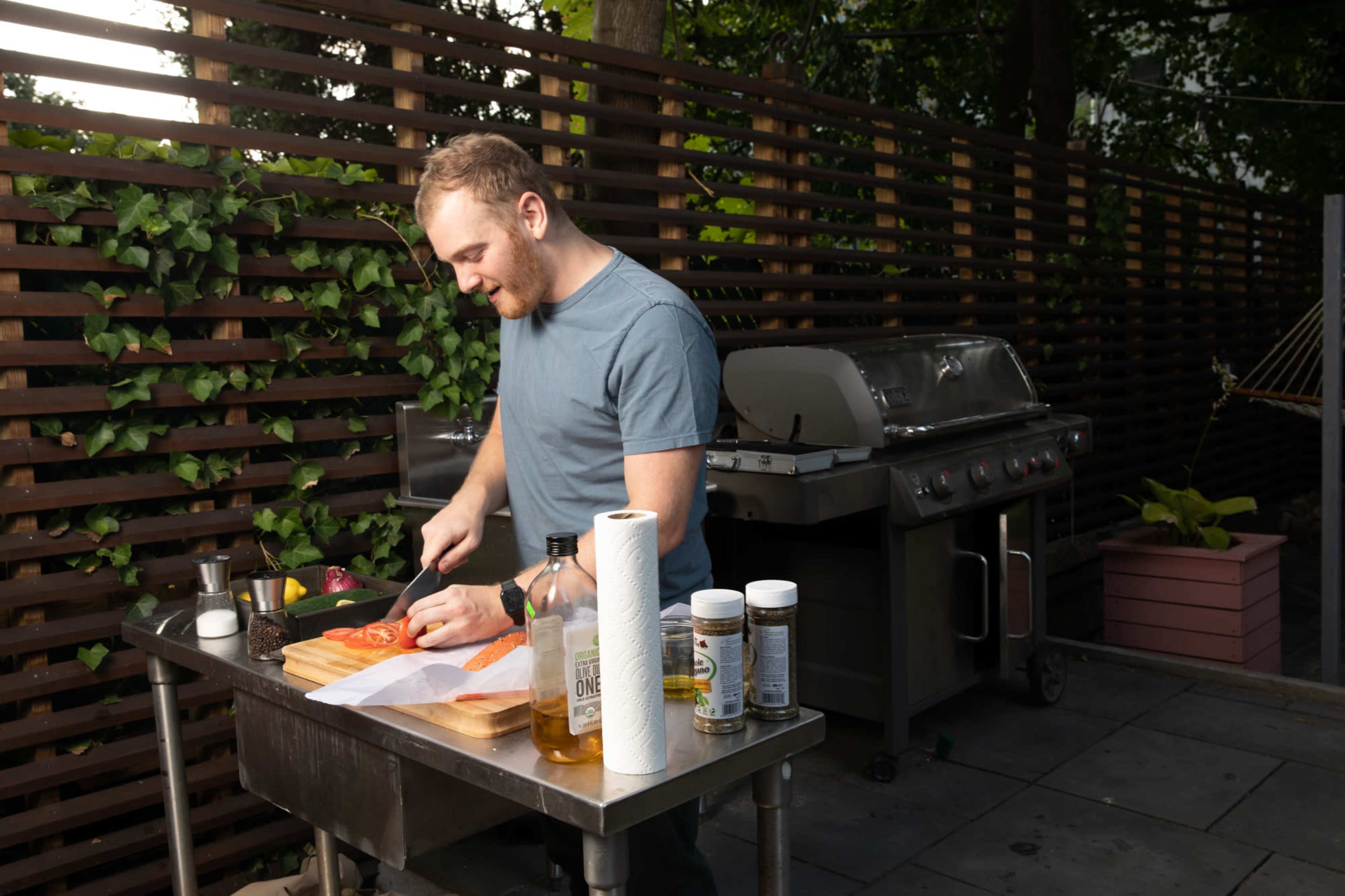 A man prepares vegetables on a wooden cutting board at an outdoor grill station surrounded by greenery.
