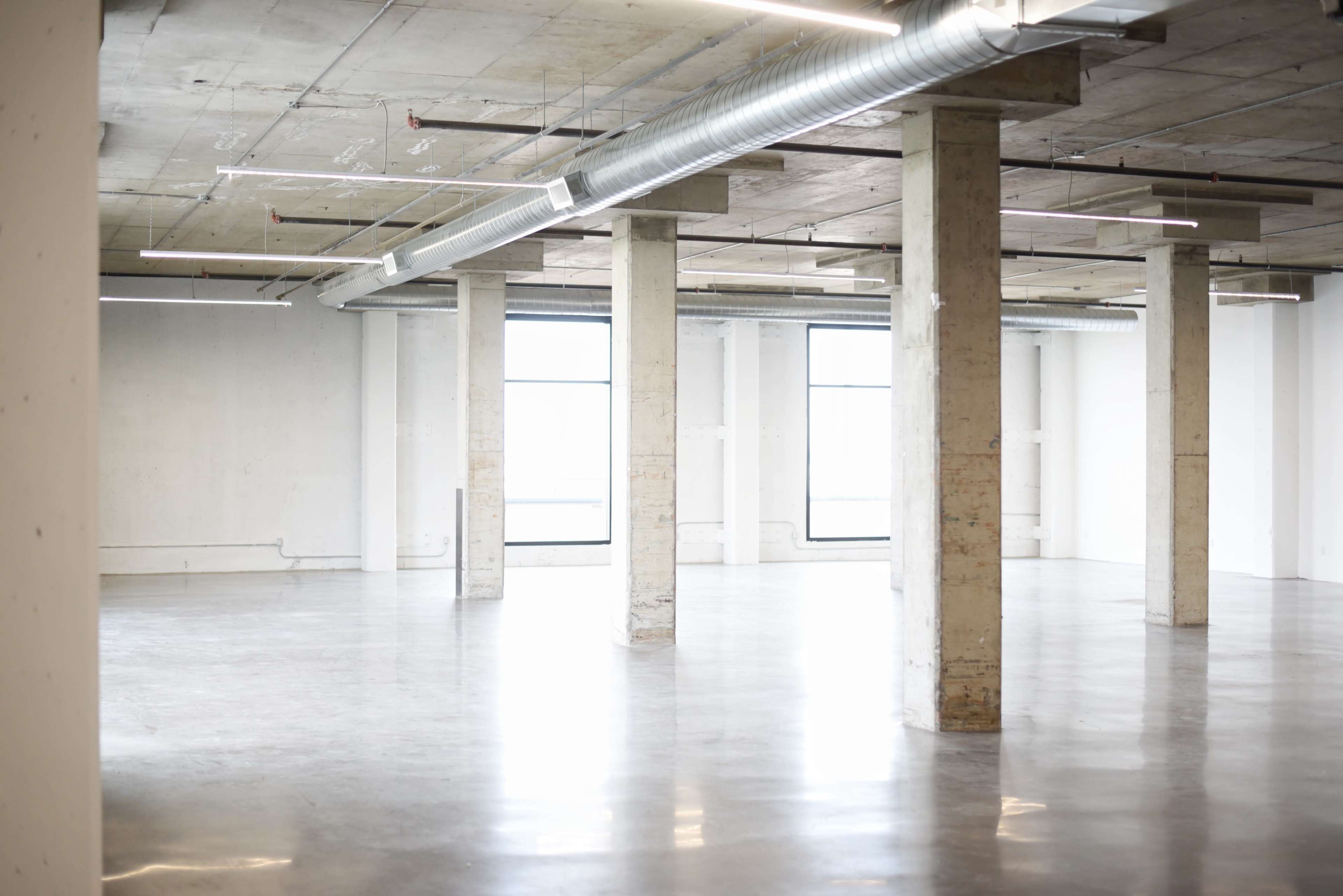 The image shows an empty, spacious interior with concrete pillars and large windows allowing natural light to enter.