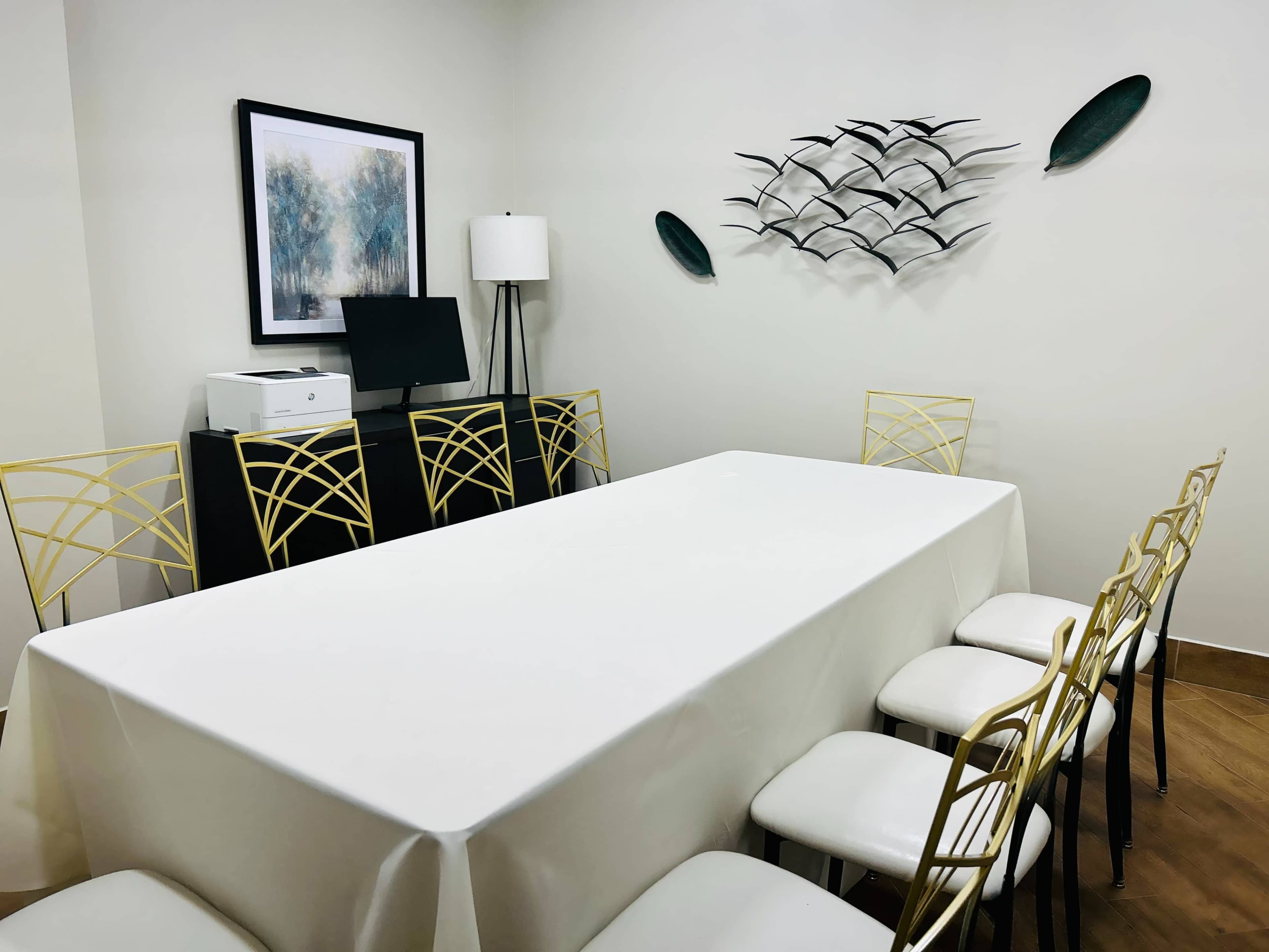 A rectangular table covered with a white tablecloth is set in a room featuring a lamp, computer, and decorative wall art.