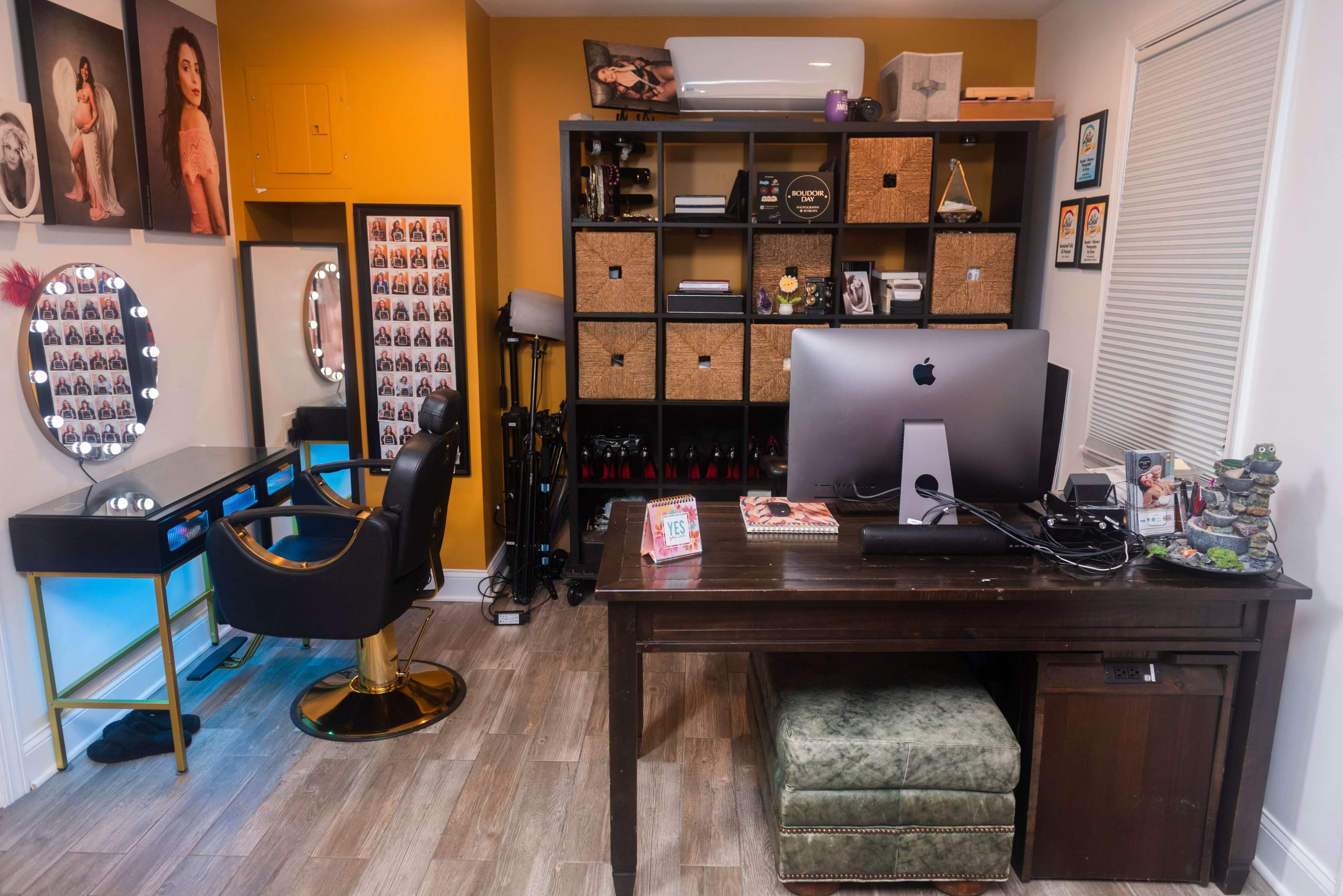 The image shows a brightly lit home office featuring a makeup station with a large mirror, a modern desk with an iMac, and shelves filled with storage boxes and framed photos.