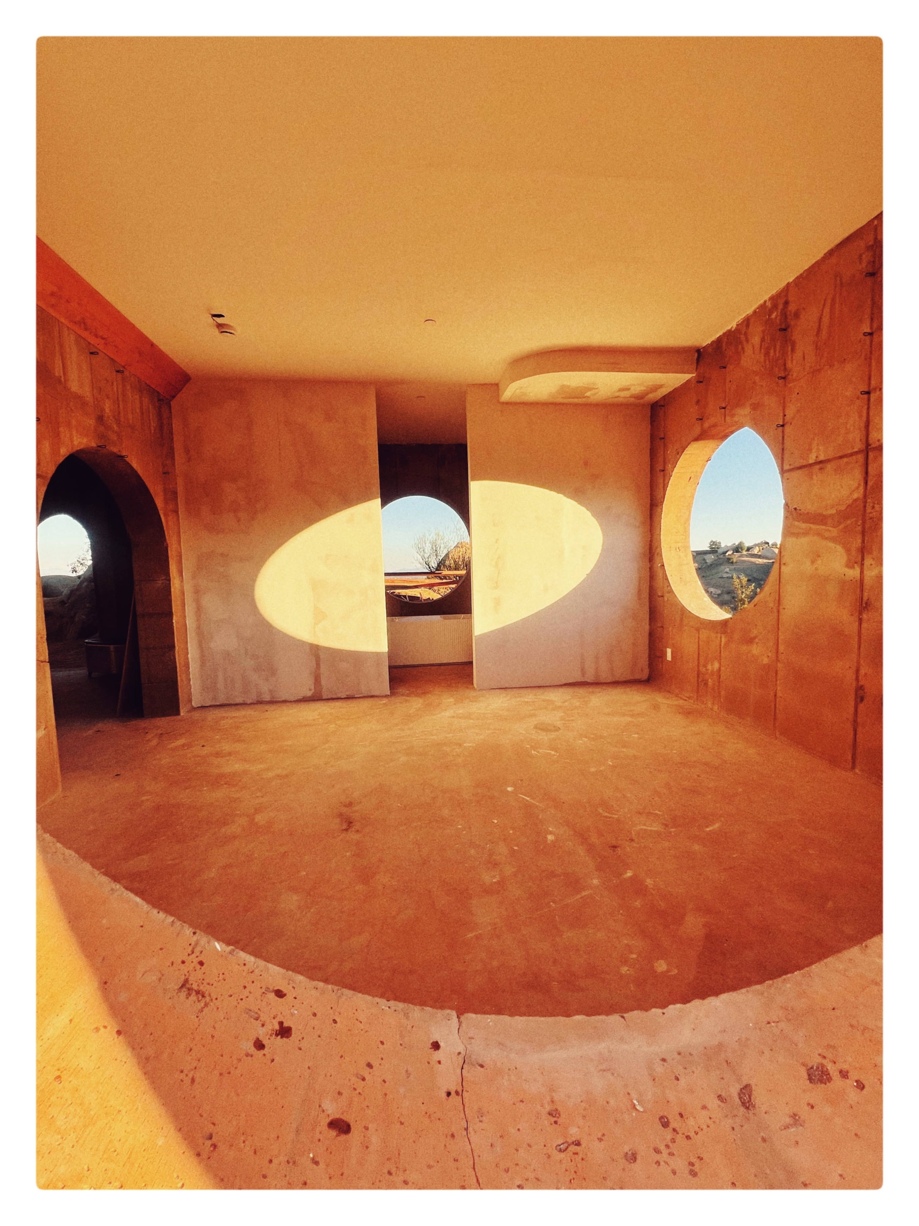 The image shows an unfinished interior space with curved walls and large circular windows allowing natural light to flow in.