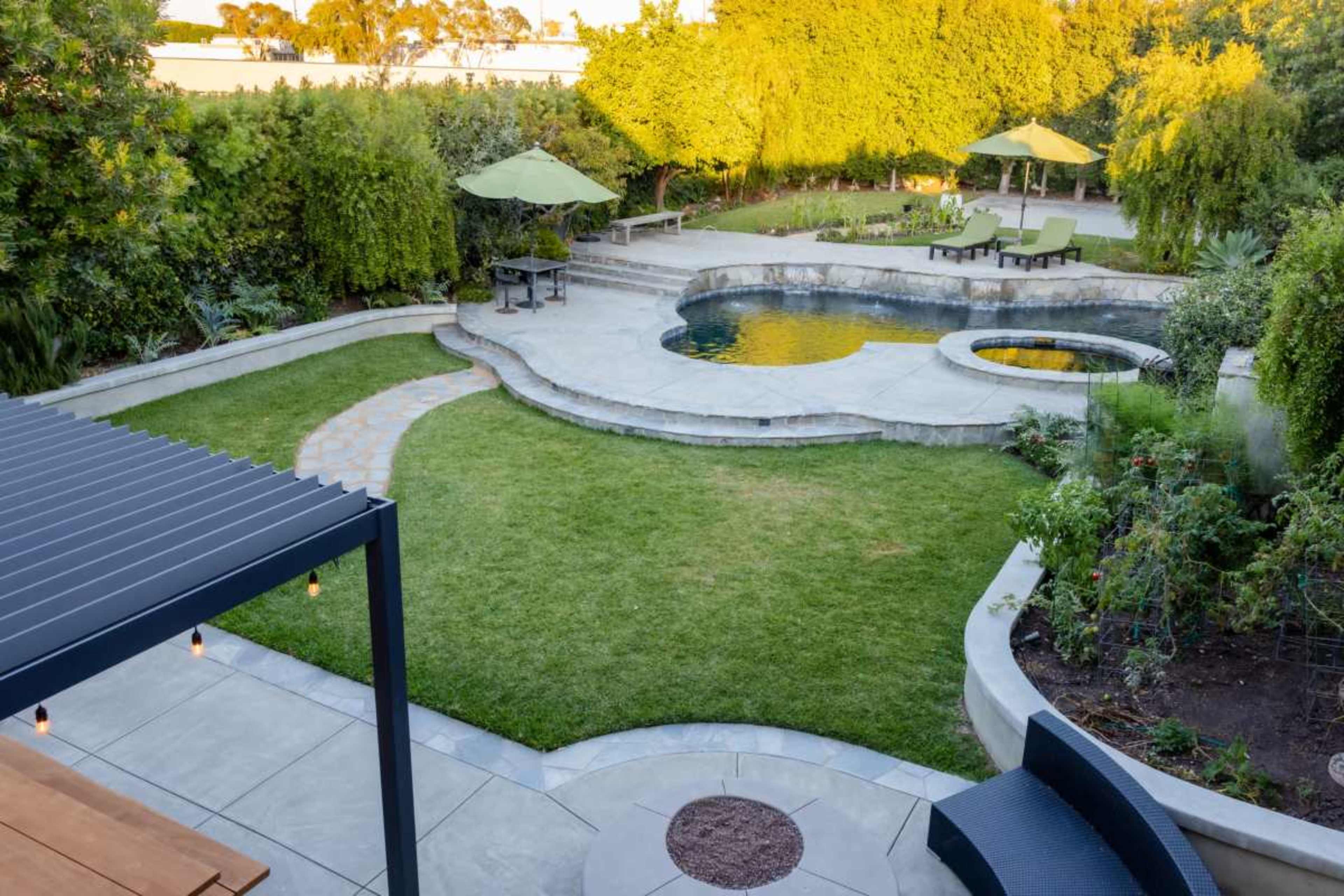 The image shows a landscaped backyard featuring a swimming pool, a circular spa, grass areas, and several seating areas under umbrellas.
