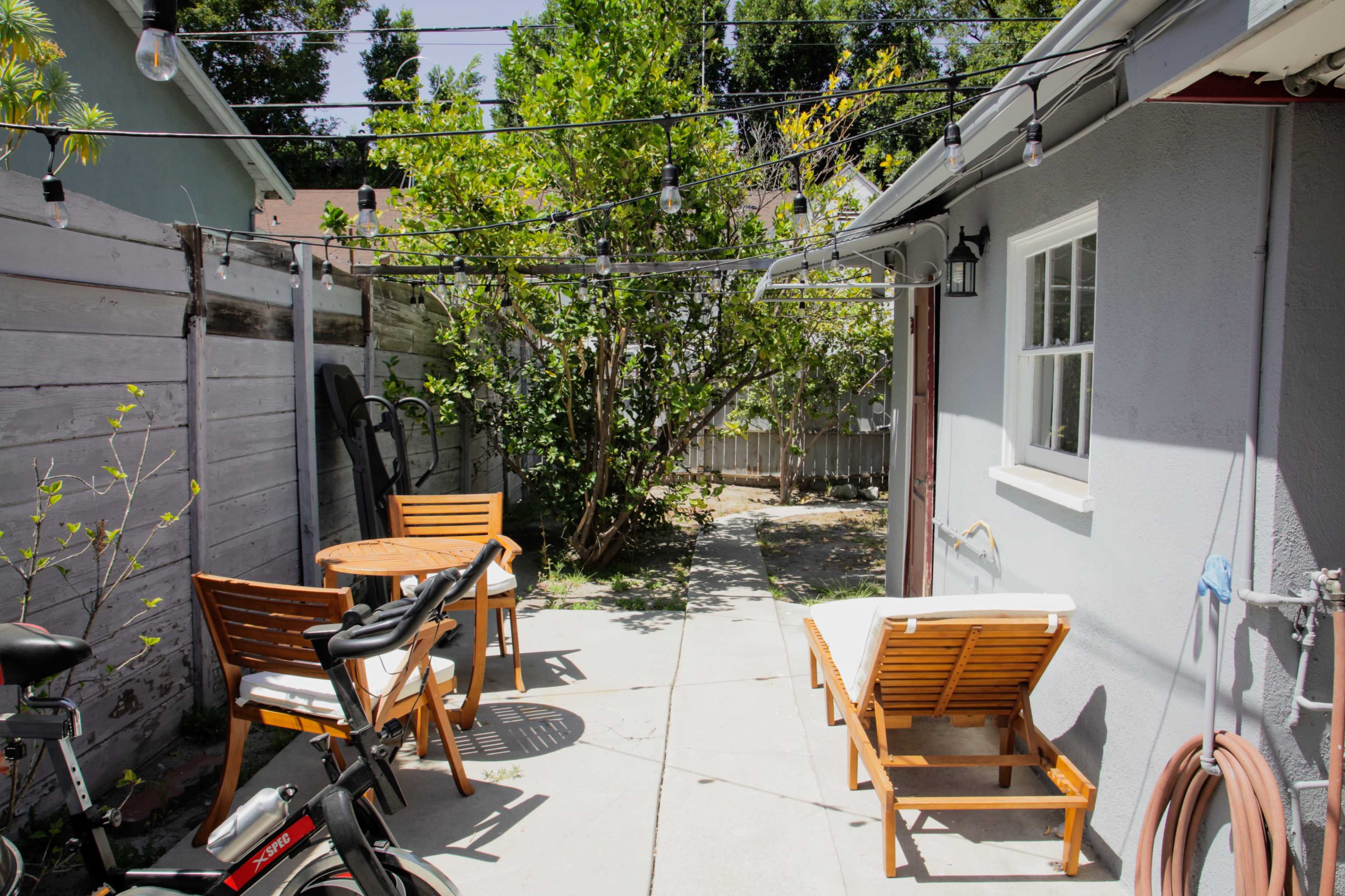 A small patio area features two wooden lounge chairs and a round table, surrounded by plants and string lights.