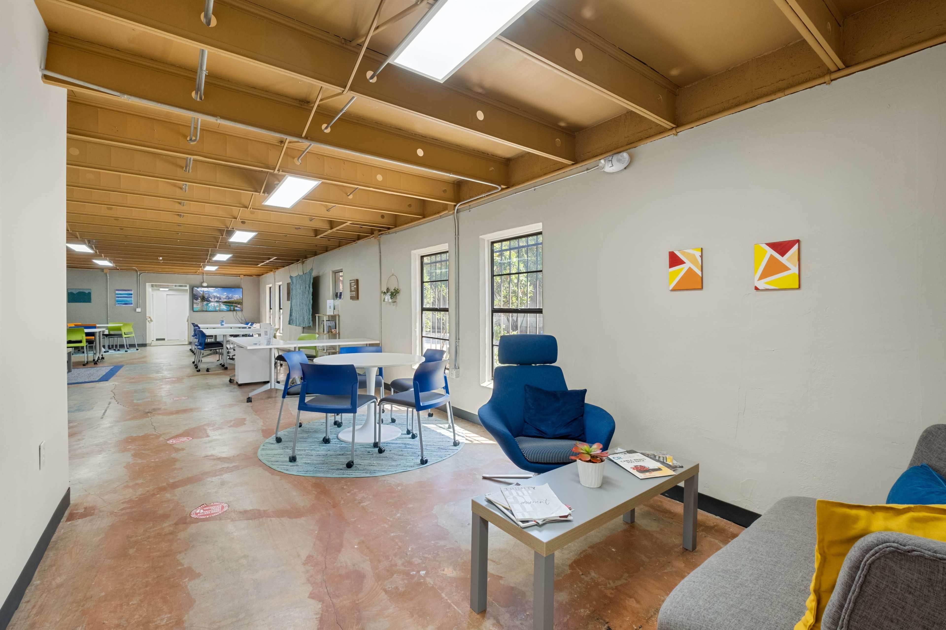 The image shows a spacious, well-lit office area with a mix of seating arrangements, including a blue chair, tables, and a gray couch along one wall.