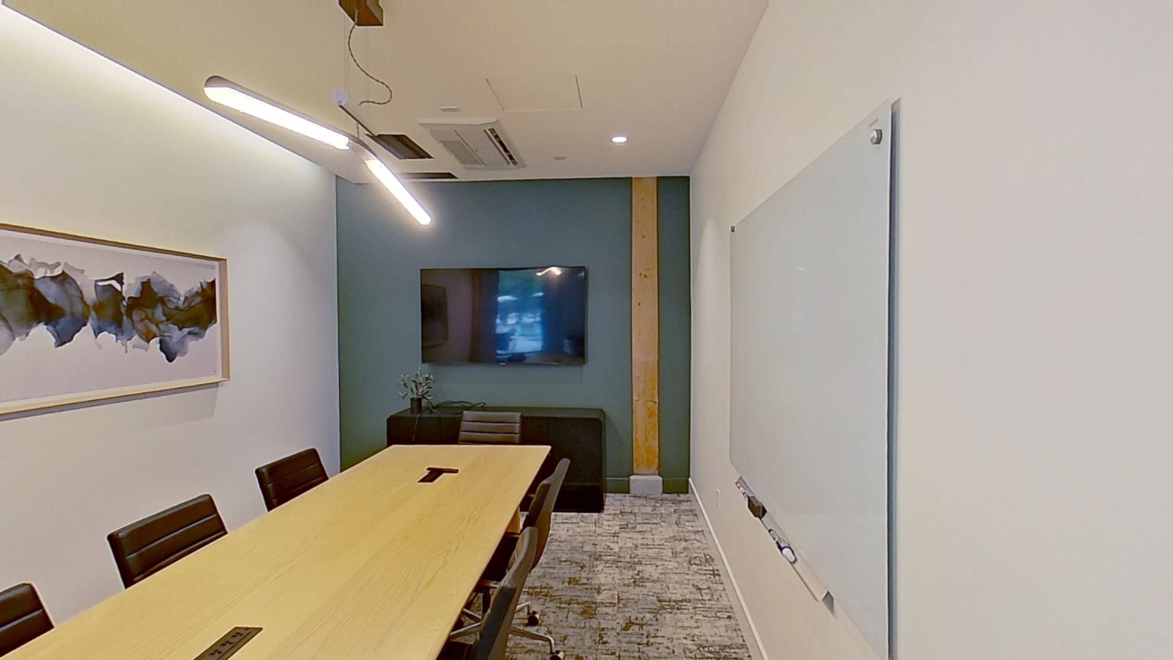 A conference room features a long wooden table, a wall-mounted television, a whiteboard, and modern lighting.