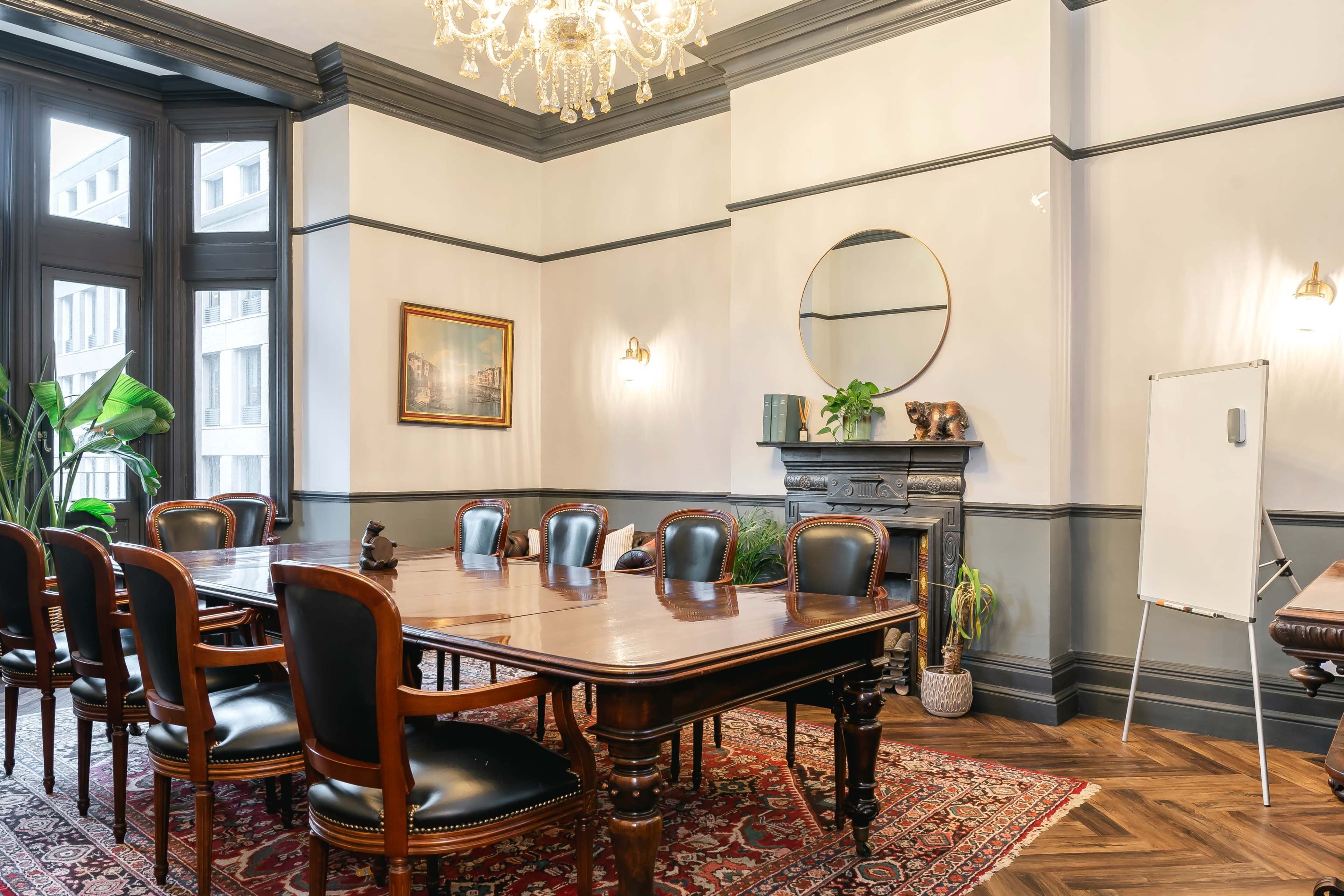 A conference room features a large, polished wooden table surrounded by leather chairs, a decorative fireplace, and a whiteboard.