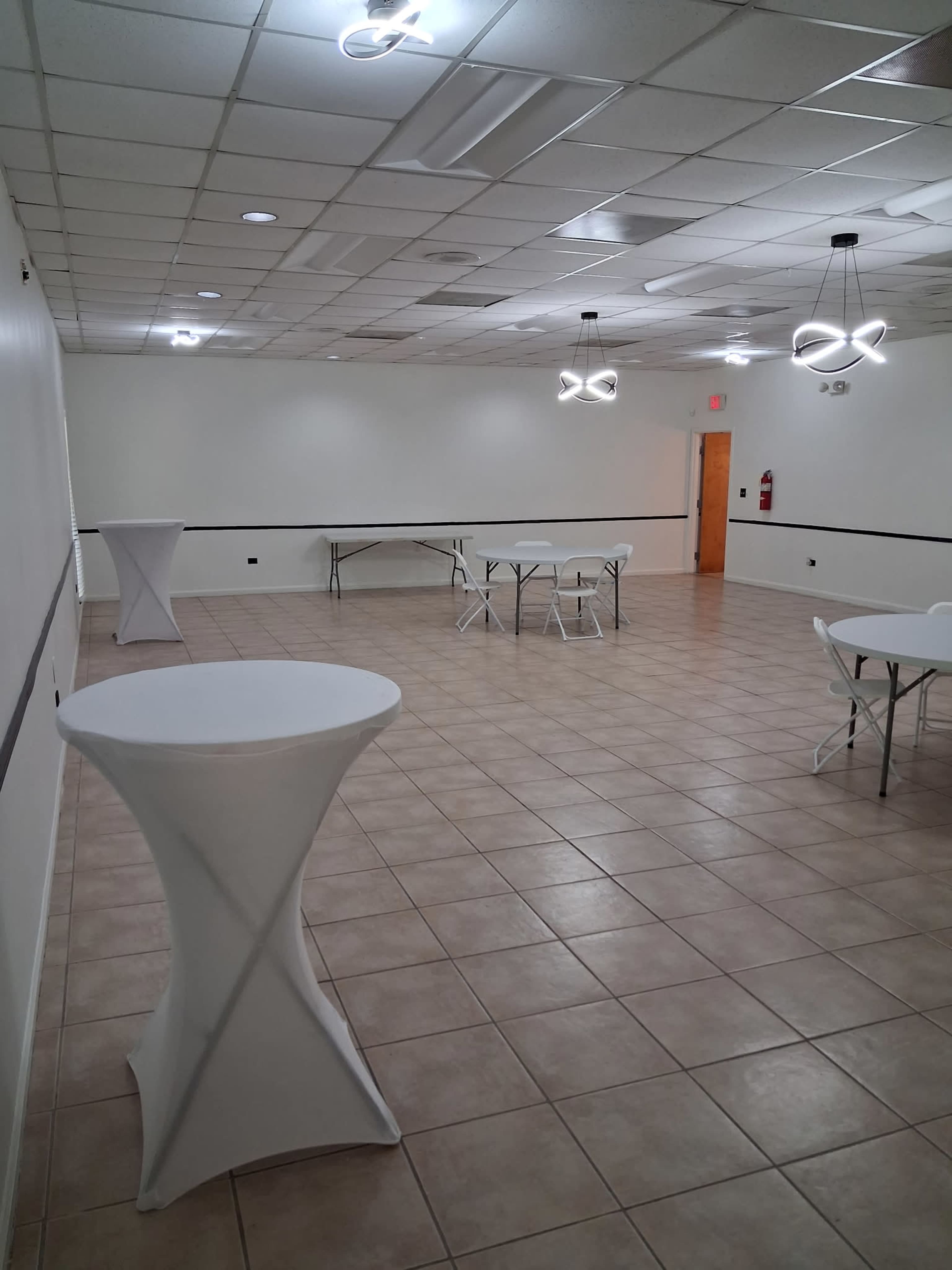 The image shows an empty event space with white walls, tiled flooring, two round tables, several tall white cocktail tables, and modern light fixtures on the ceiling.