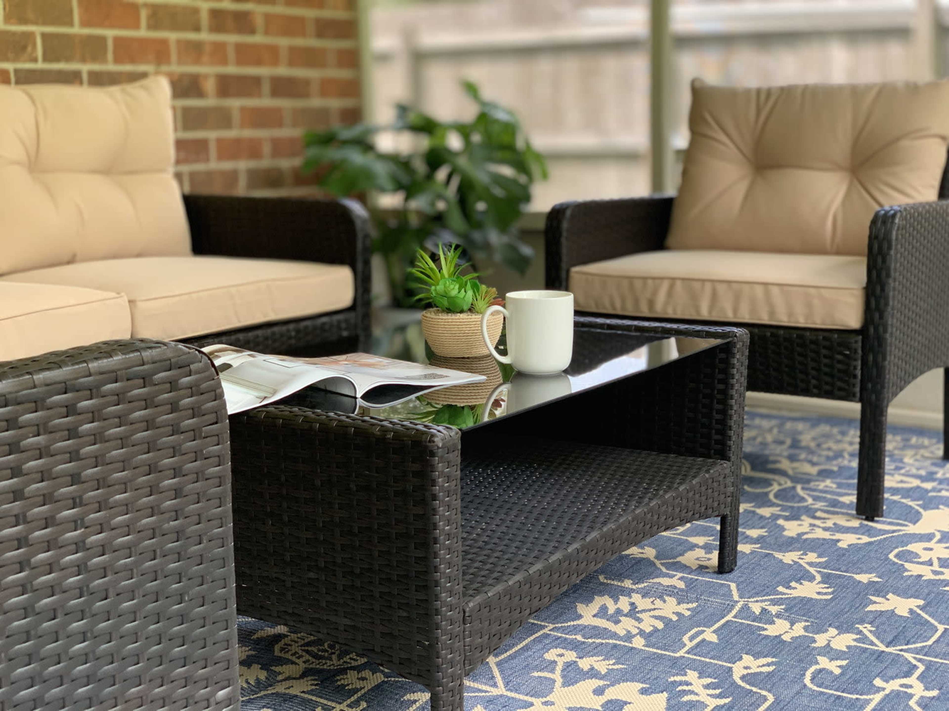 A cozy seating area with two wicker chairs and a coffee table, set on a patterned rug, with a plant and a magazine on the table.