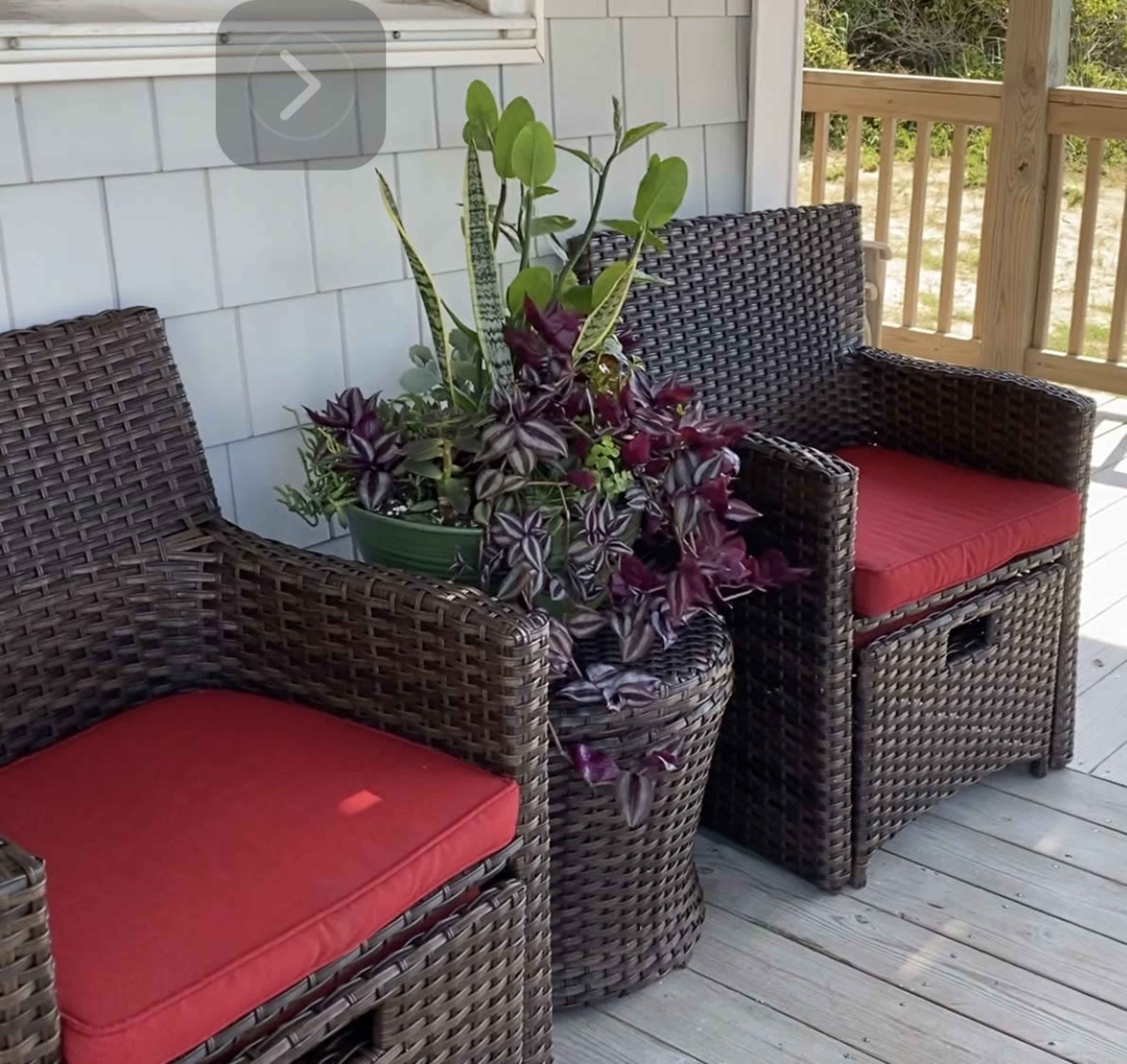 Two wicker chairs with red cushions sit adjacent to a planter filled with various green and purple plants on a wooden porch.