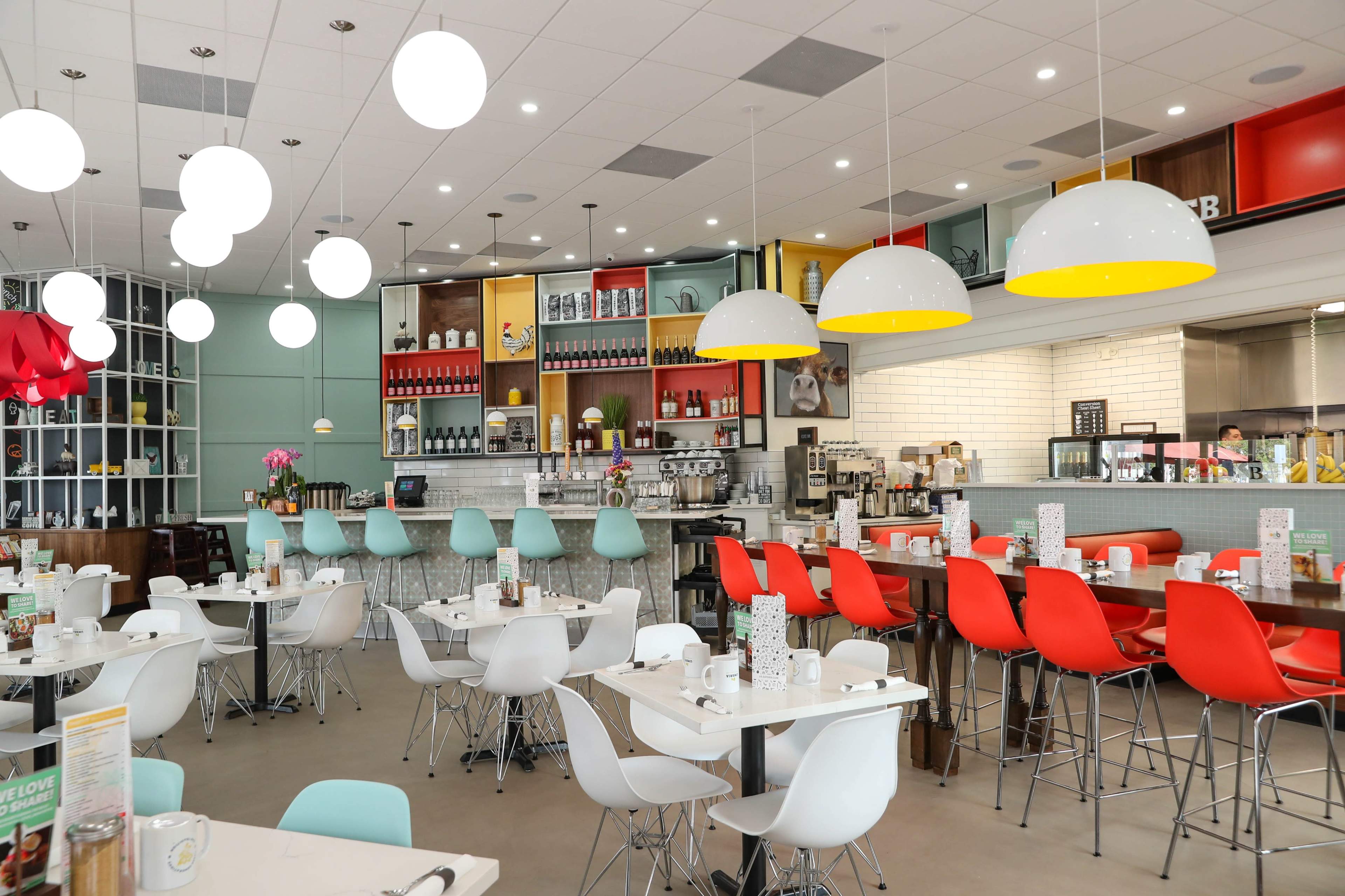 A modern restaurant features colorful chairs and pendant lighting, with tables set for dining and a well-stocked bar area in the background.