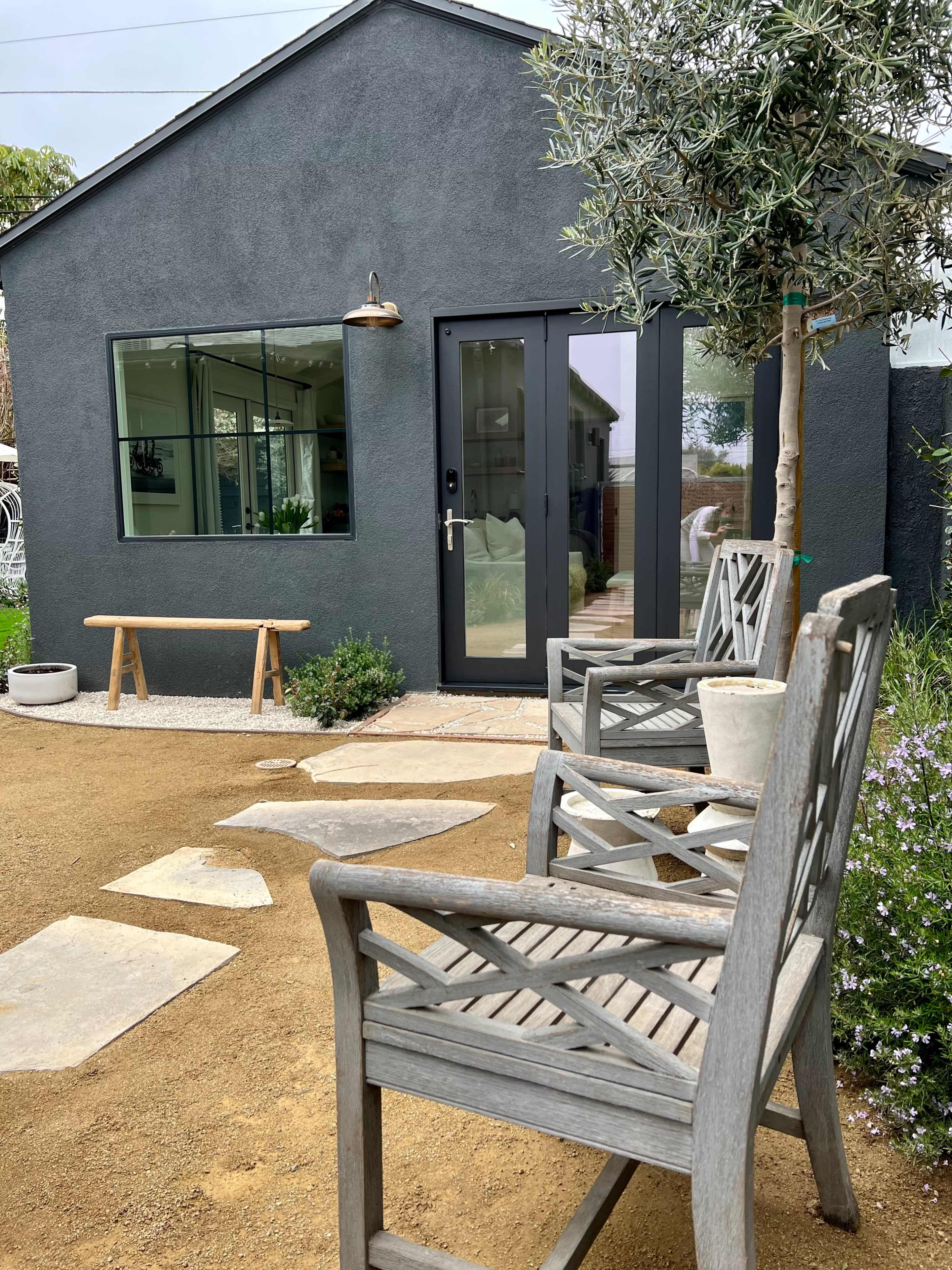 A modern dark-colored house with a large glass door, surrounded by gravel and plants, features wooden seating in front.