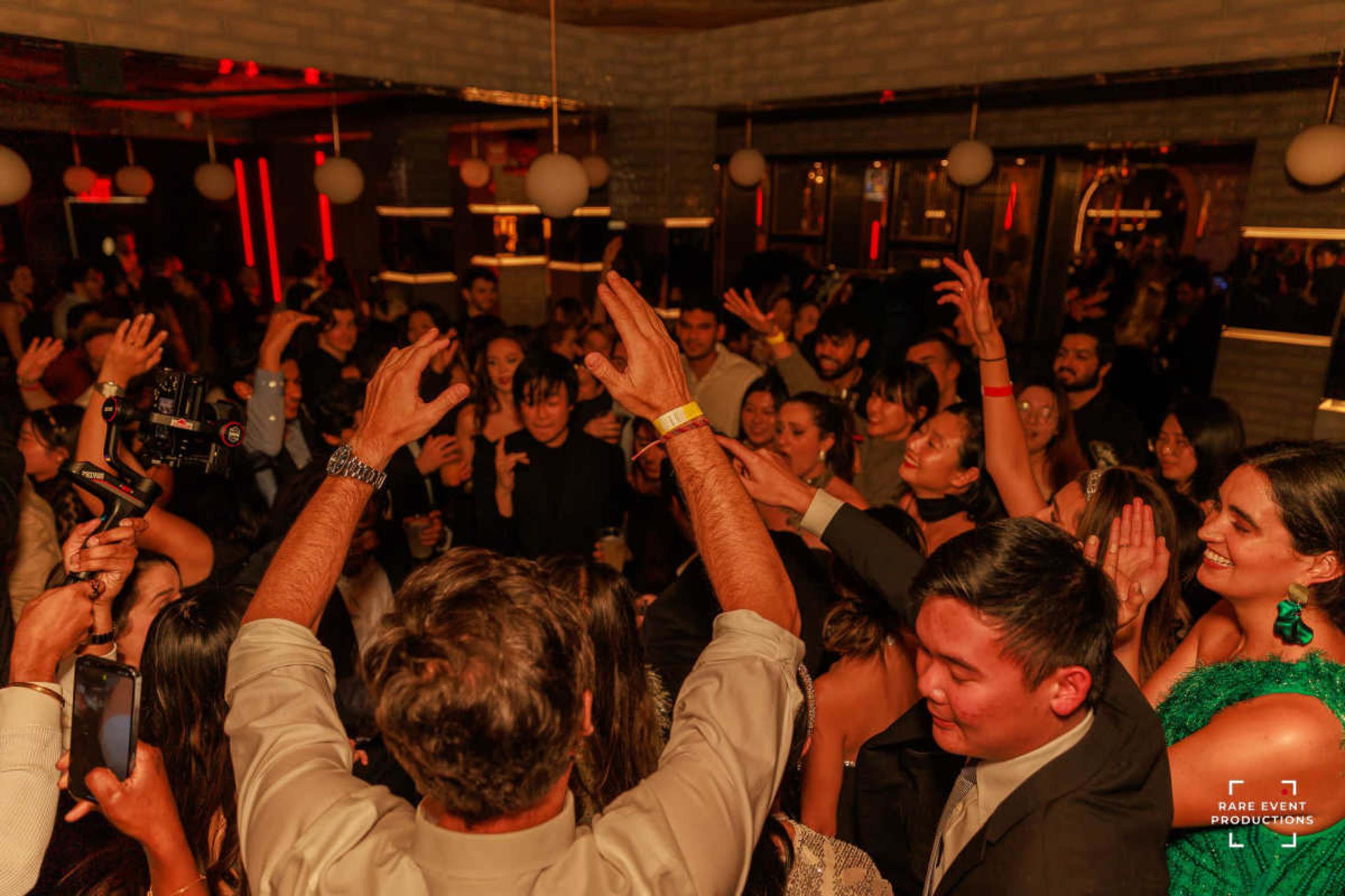 A large crowd is dancing enthusiastically in a dimly lit venue with vibrant lighting.