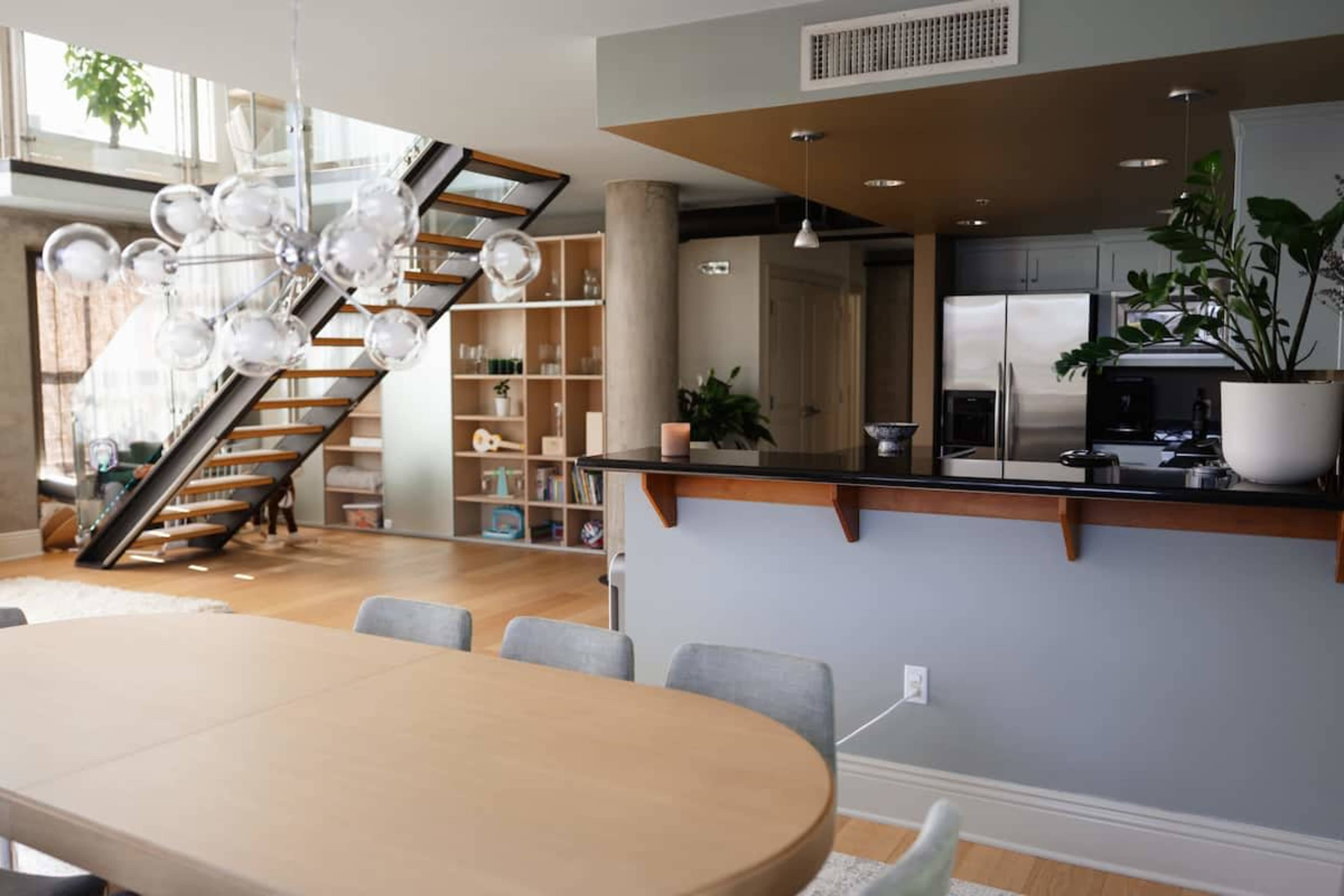 The image depicts a modern interior space featuring a dining area with a wooden table, a staircase in the background, and a kitchen equipped with stainless steel appliances.