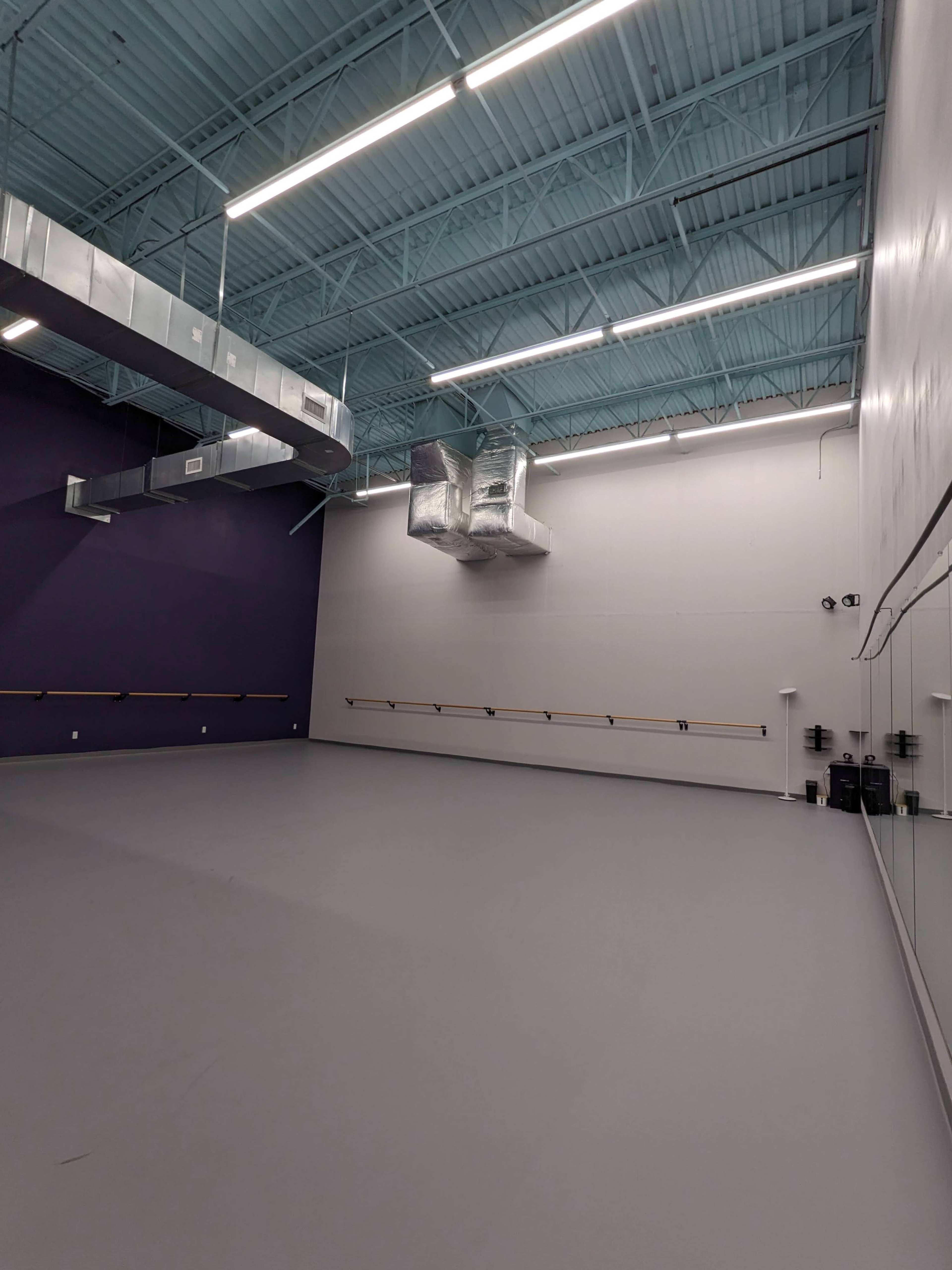The image shows a spacious dance studio with a gray floor, mirrors on one wall, and a high ceiling painted blue.