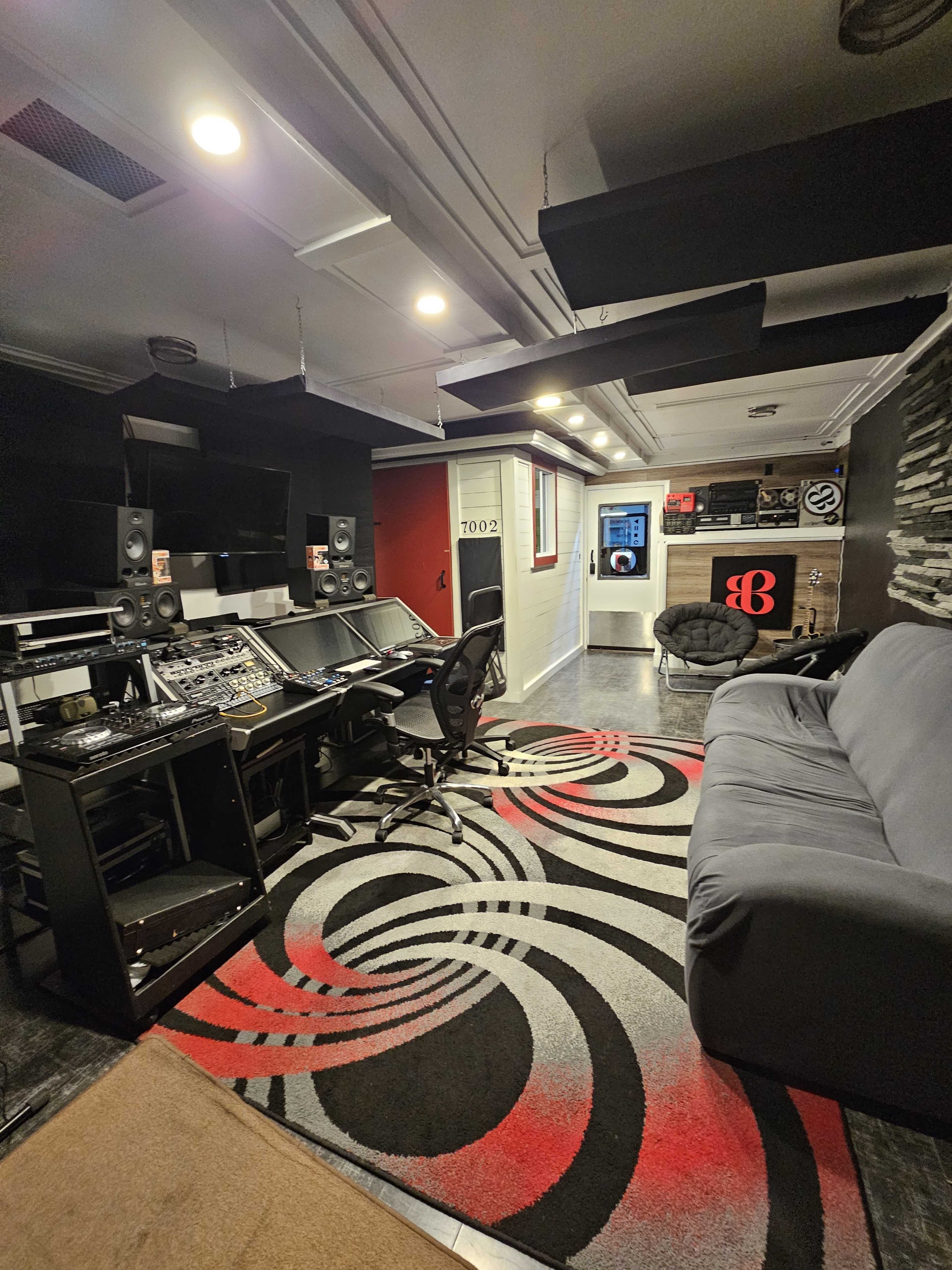 The image features a modern recording studio with black walls, a sound mixing console, computer monitors, and a large rug with a spiral design, alongside a sofa and a cozy chair.
