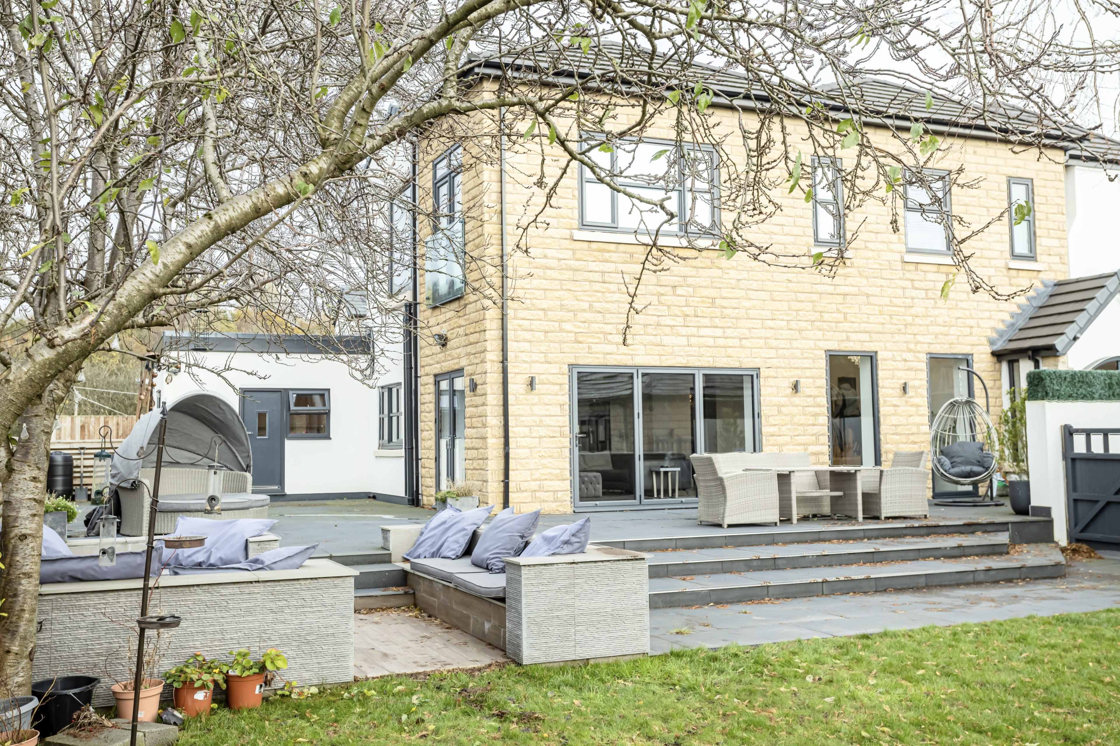 The image shows a modern two-story house with stone exterior and large glass doors leading to a patio area featuring outdoor seating and a hanging chair.