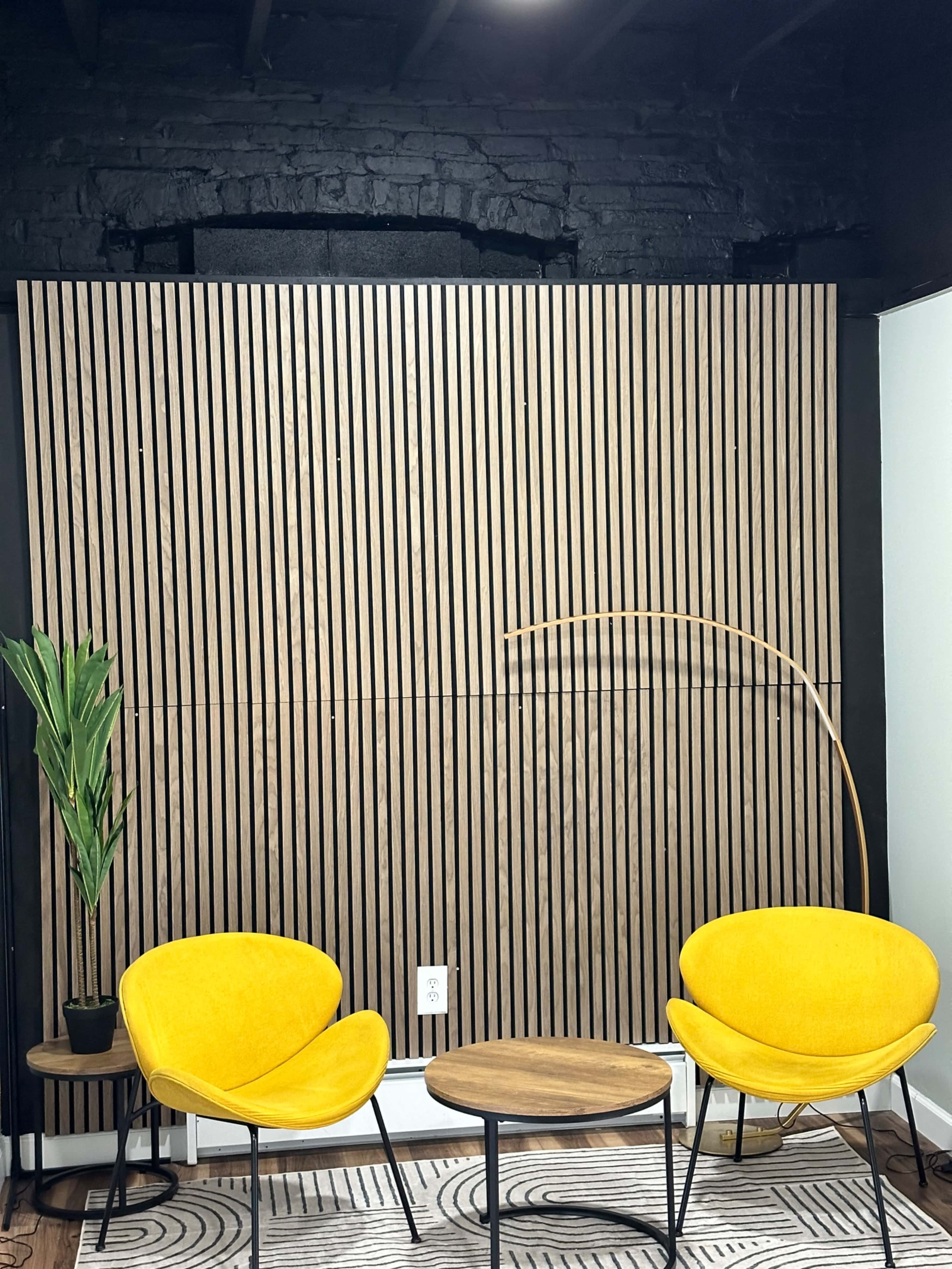 The image shows a minimalist interior featuring two yellow chairs, a round wooden table, a potted plant, and a wall with vertical wooden slats.
