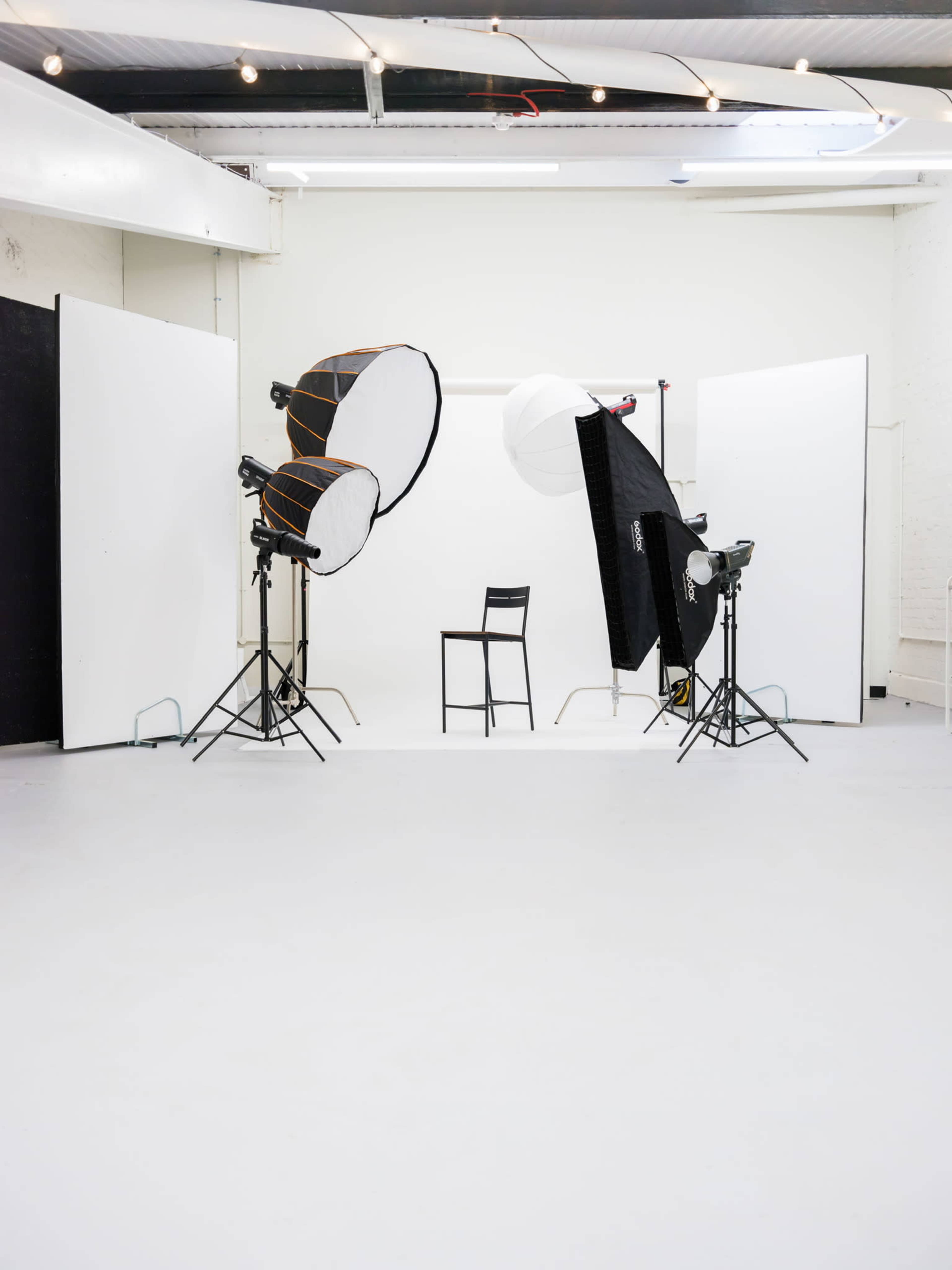 A photography studio is set up with two lighting setups and a single chair positioned in the center against a white backdrop.