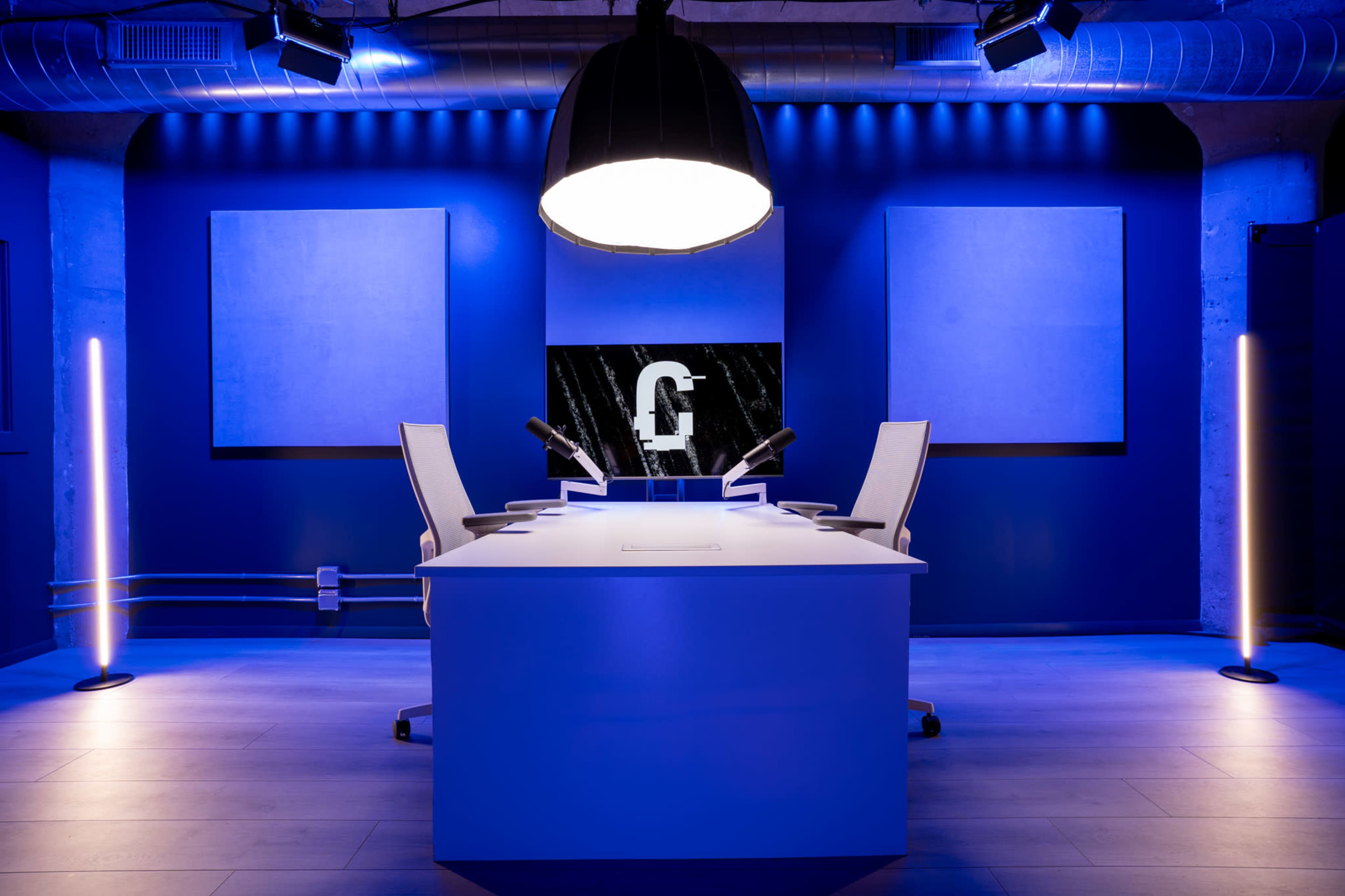 A modern studio setting features a central white table with two chairs, illuminated by a large overhead light and accented by blue wall lights, against a backdrop of dark blue walls and empty framed canvases.