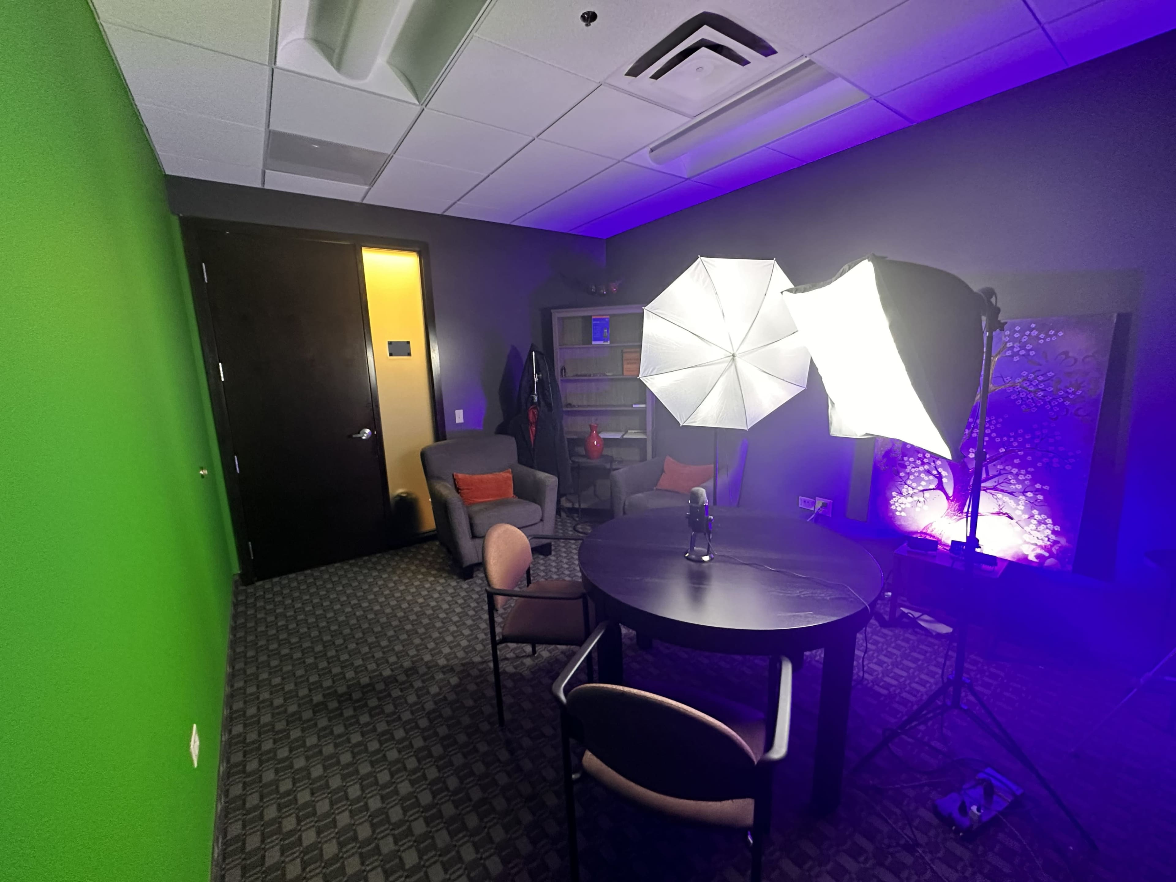 A small room features a green screen wall, two lighting setups, a round table, and two chairs arranged around it.