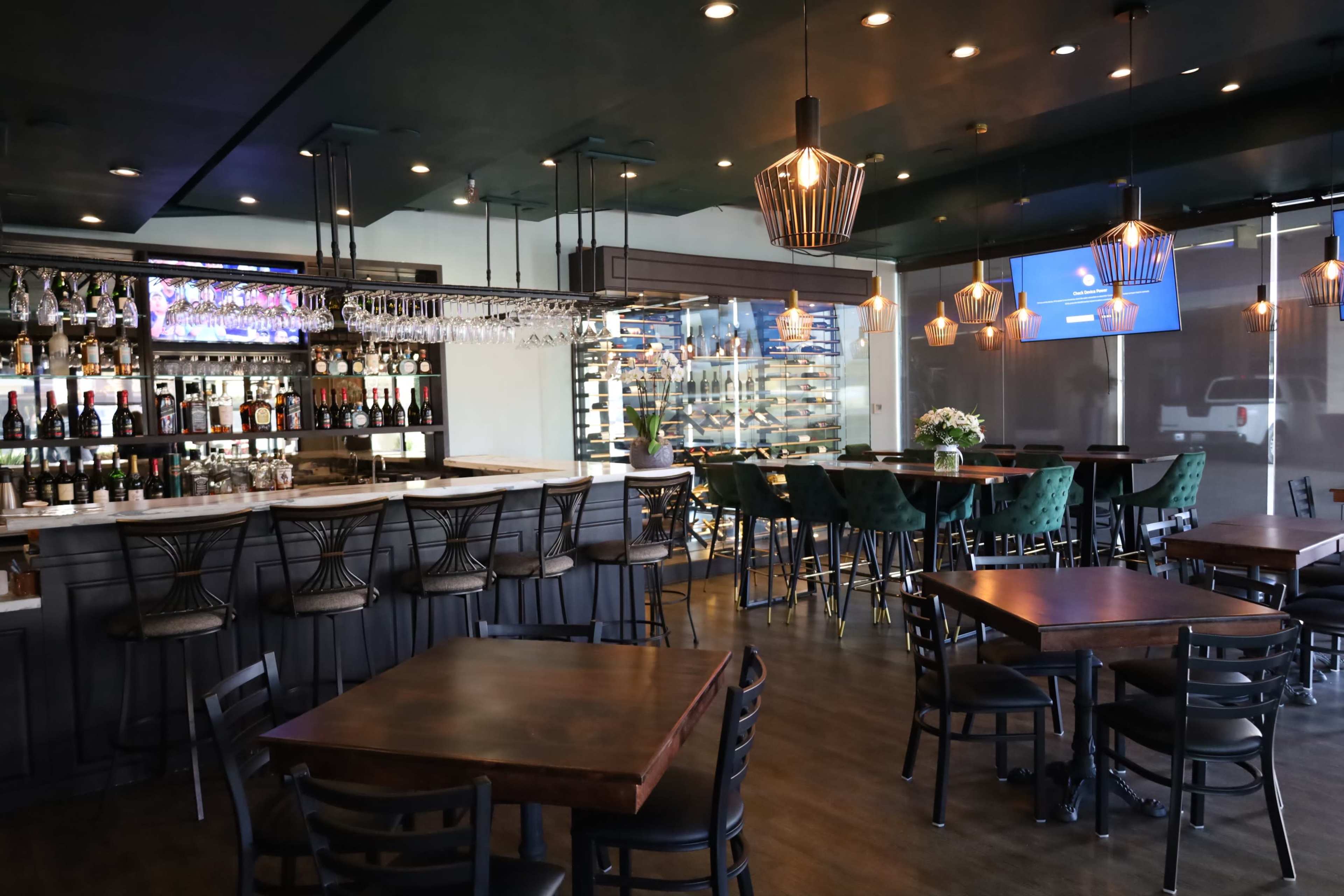 The image shows a modern bar and dining area with dark furnishings, a well-stocked liquor display, and ambient lighting.