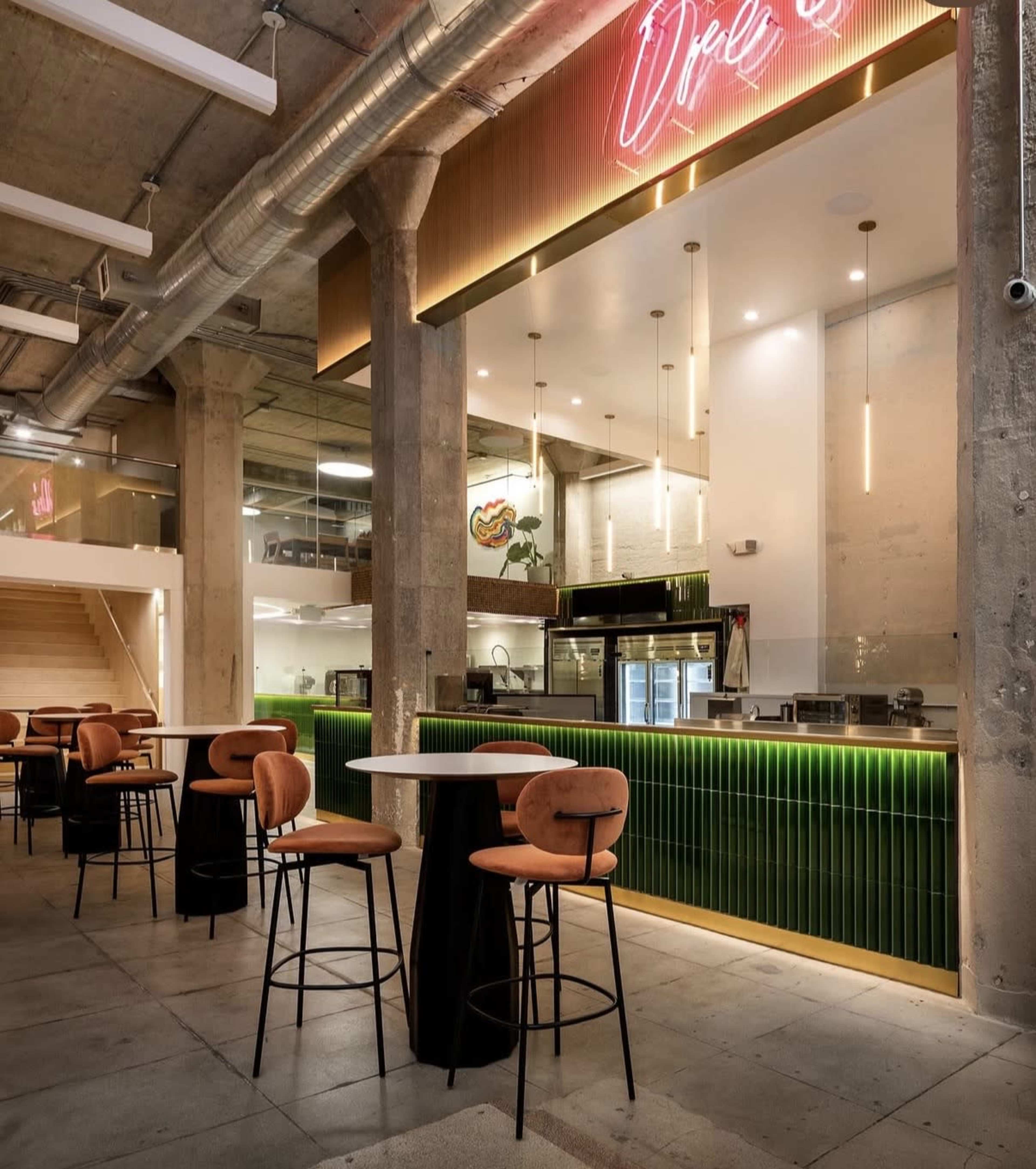 The image shows a modern café interior featuring a neon sign, a green tiled counter, high tables with stools, and an industrial-style ceiling with exposed pipes.