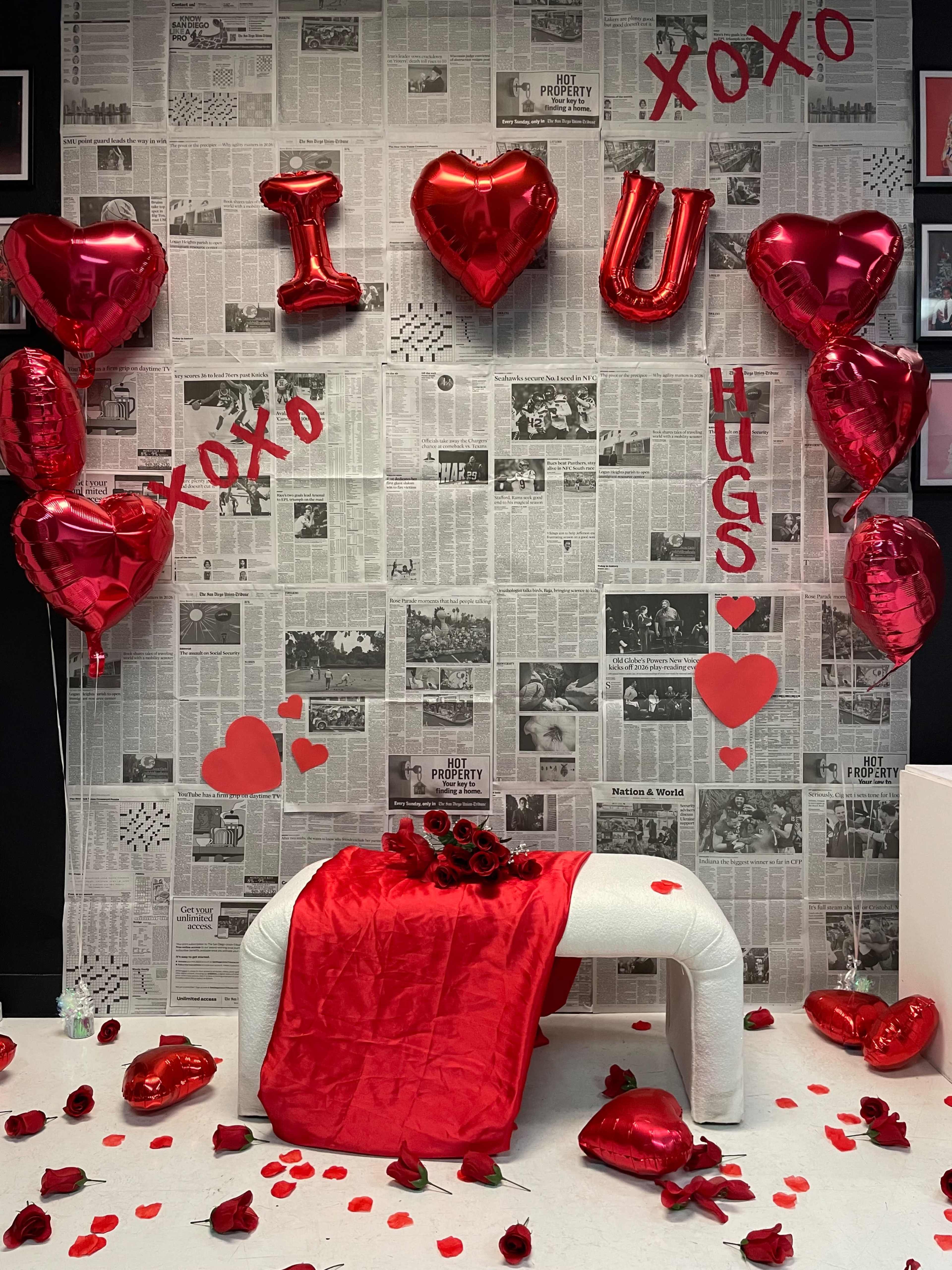 The scene features a backdrop of newspaper clippings adorned with large red balloons shaped like hearts, the letters "I," "U," and phrases like "HUGS" and "XOXO," along with a red blanket on a white bench surrounded by rose petals.
