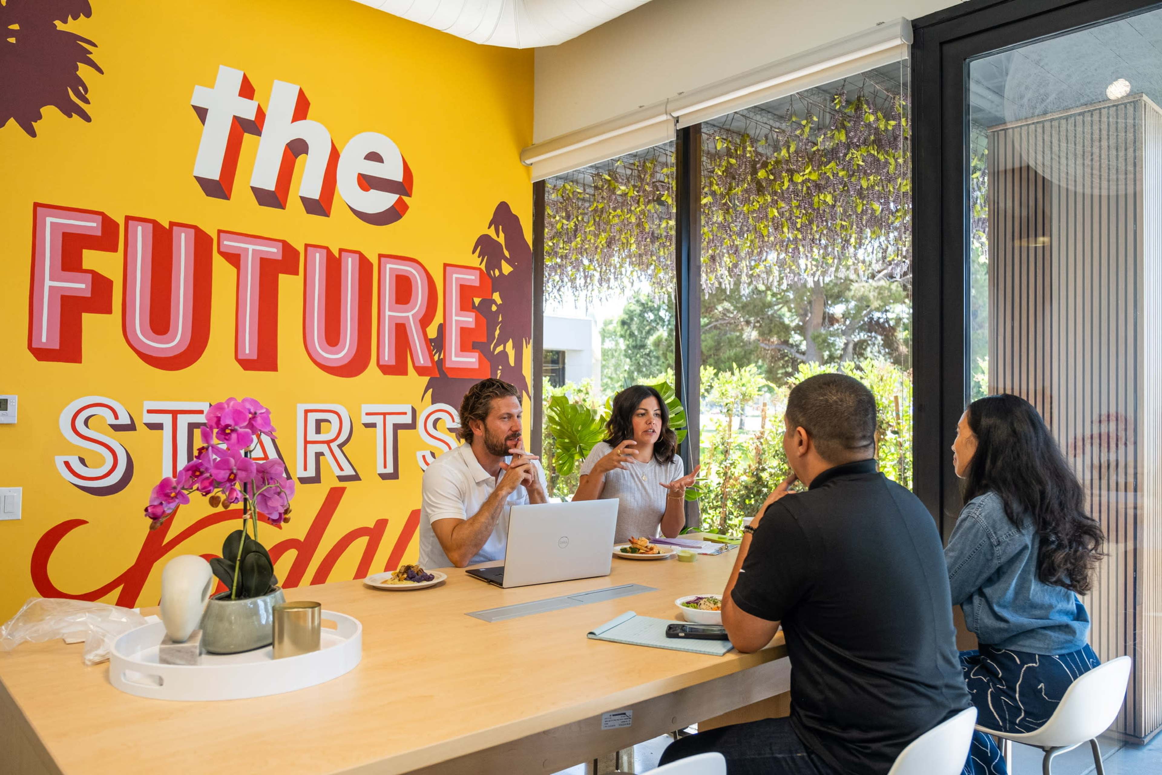 A group of four people sits around a table in a brightly colored meeting room with the phrase "the FUTURE STARTS today" on the wall, enjoying food and discussing.