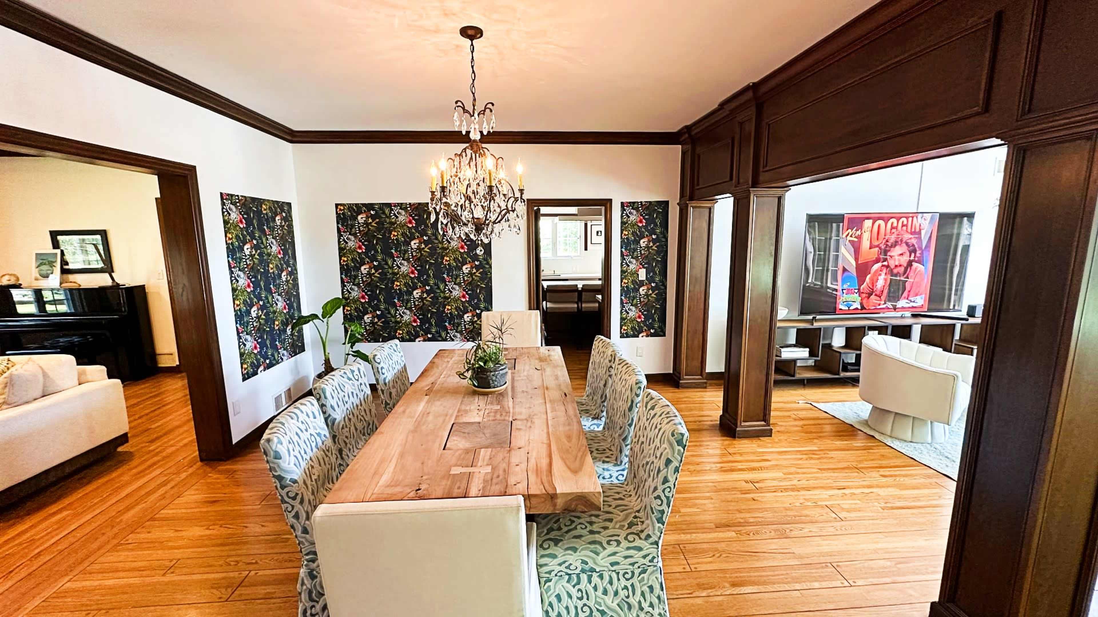 A dining room features a long wooden table surrounded by patterned chairs, with floral wallpaper on the walls and a chandelier overhead.