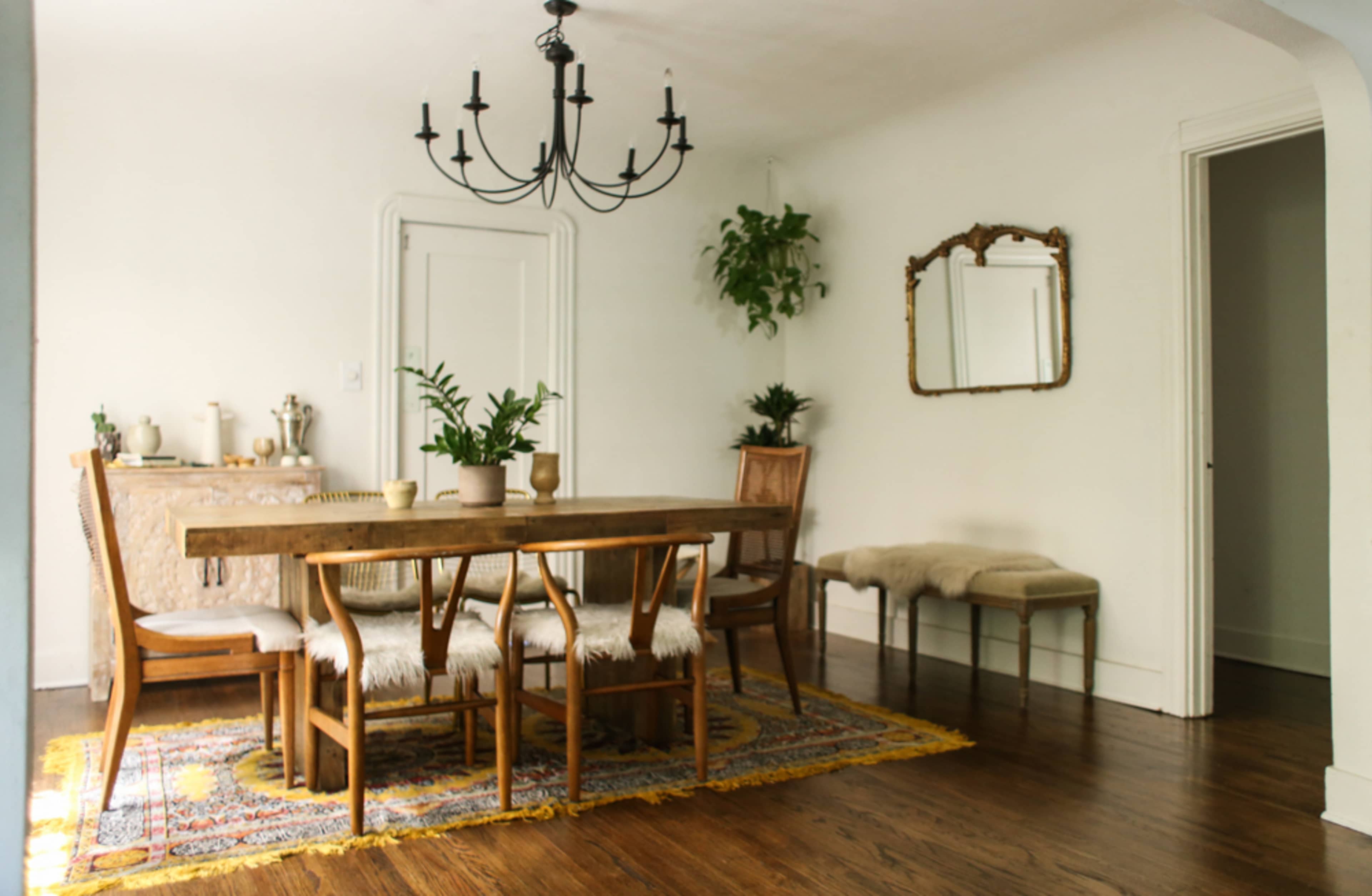 A dining room features a large wooden table surrounded by chairs, accented by a chandelier and a mirror on the wall, with potted plants adding greenery.