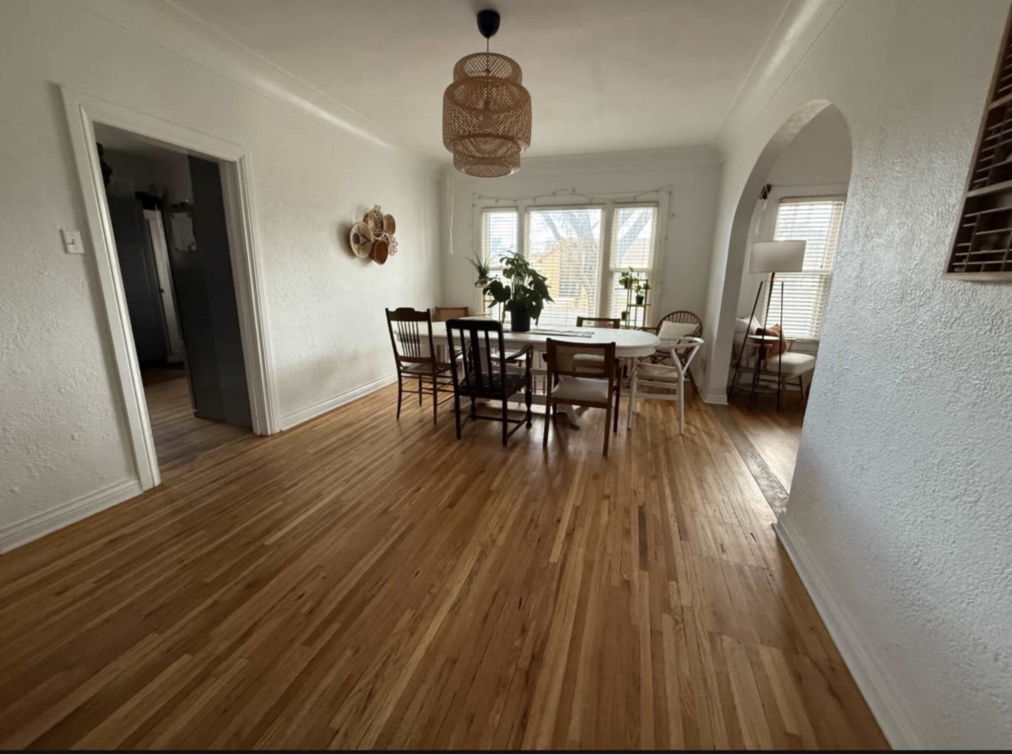 The image shows a dining area with a wooden table surrounded by chairs, hardwood flooring, and large windows allowing natural light to enter the space.