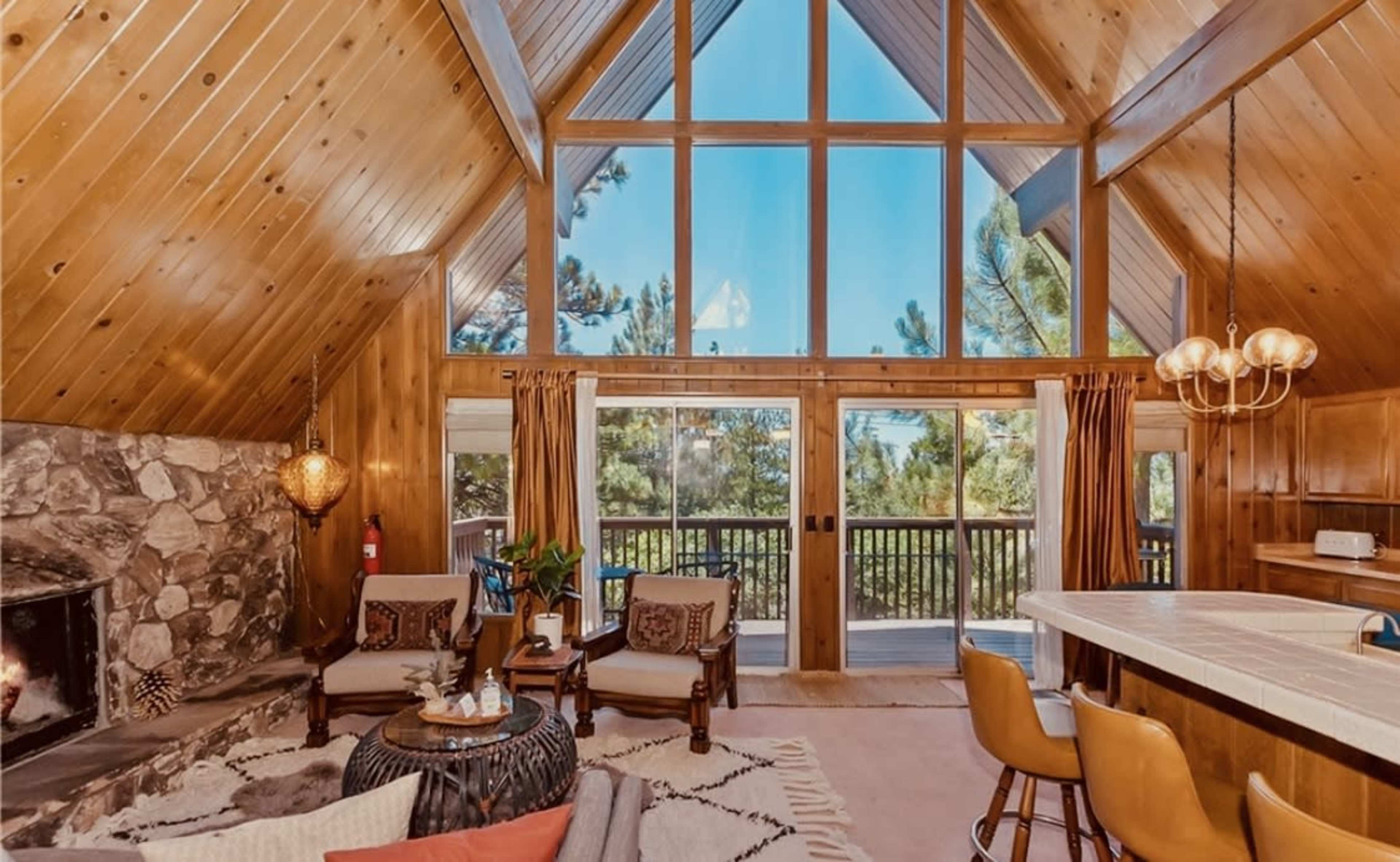 A rustic cabin interior features wooden paneling, large windows, and a stone fireplace with seating arrangements and a dining area.