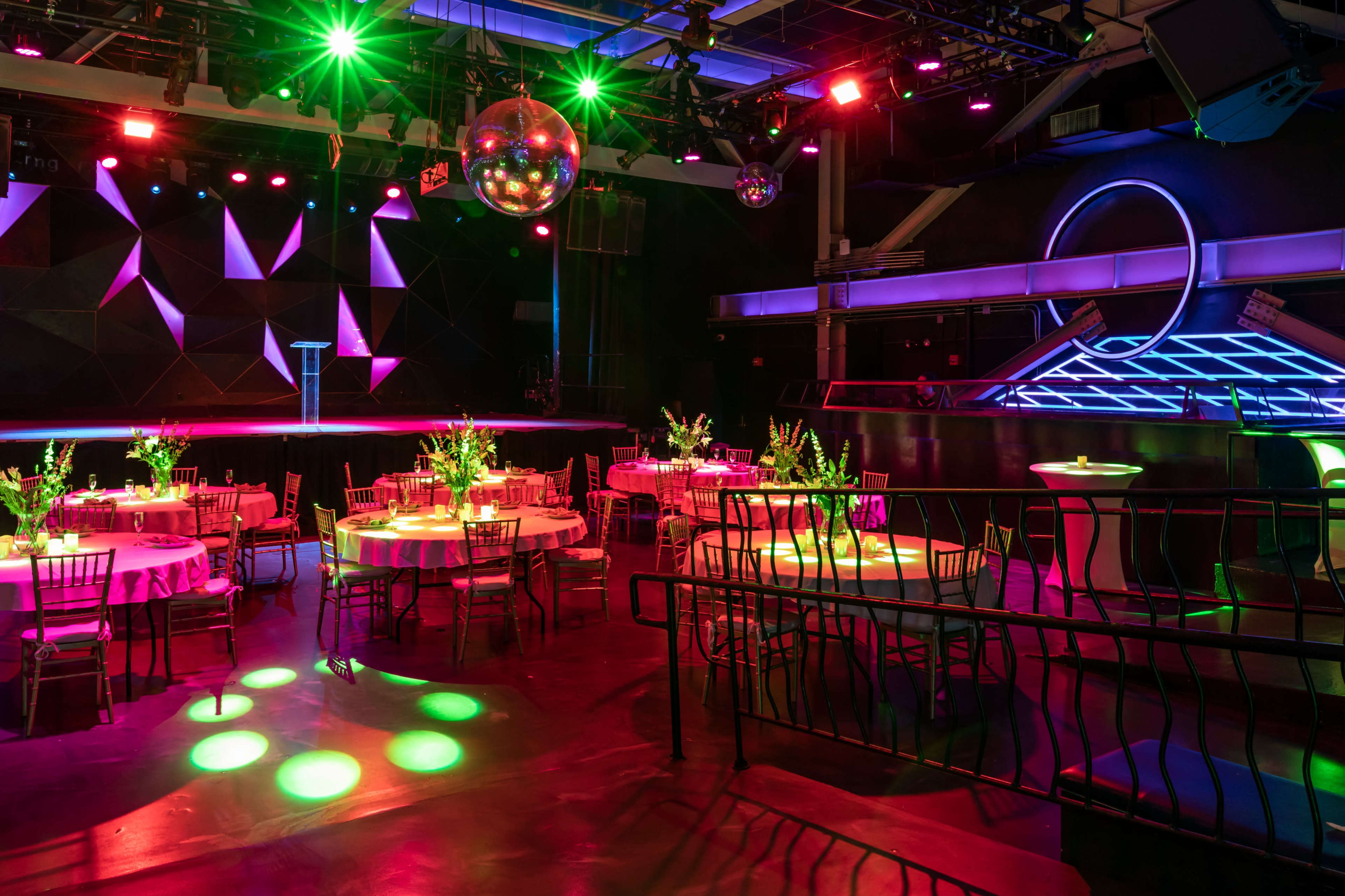 The scene displays a vibrant event space with round tables set for dining, a colorful stage illuminated with lights, and modern decor featuring a disco ball.