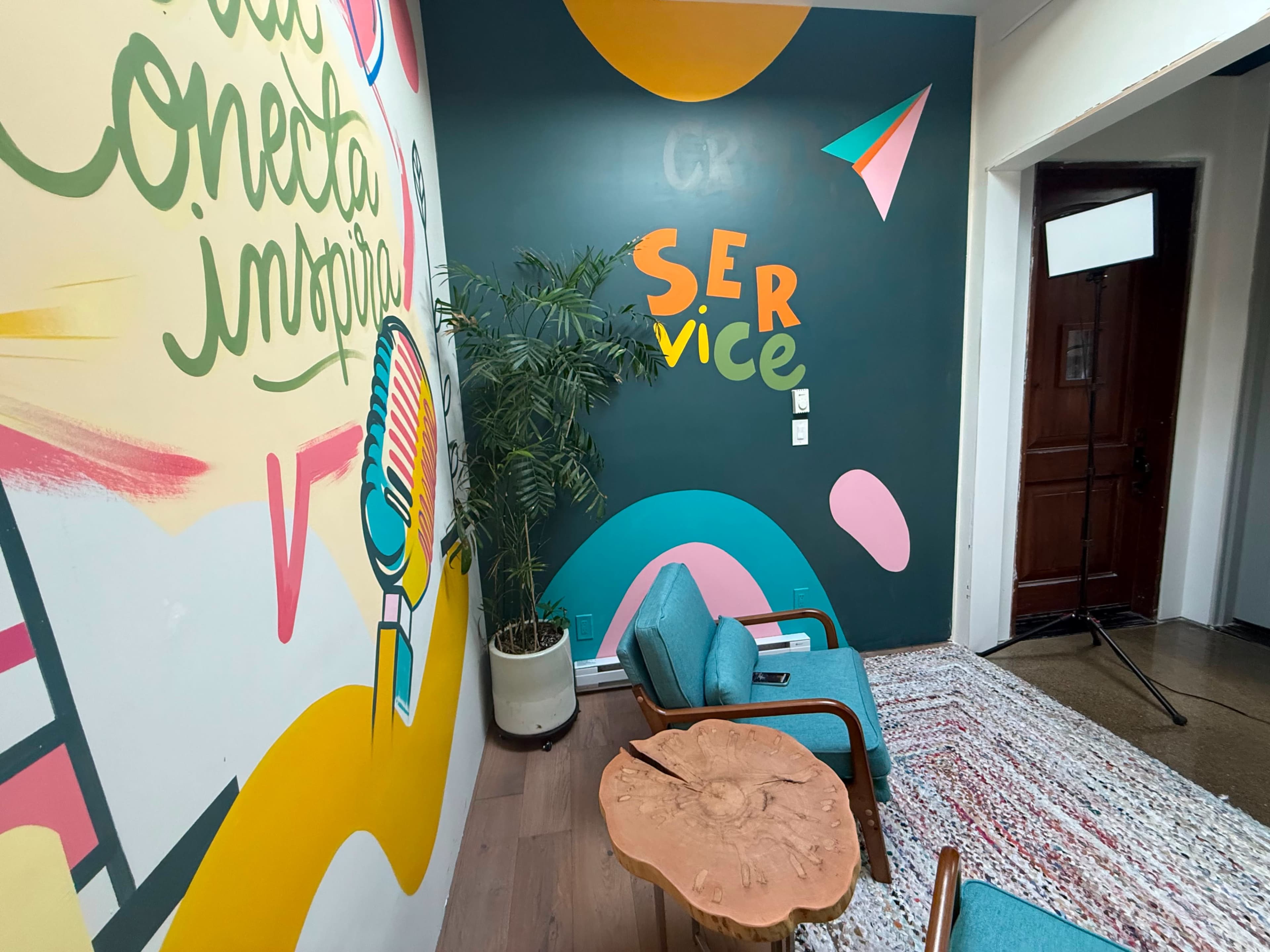 The image shows a brightly colored interior space with a mural featuring the words "Conecta Imagina" and "Service," along with a small seating area that includes turquoise chairs and a wooden table.