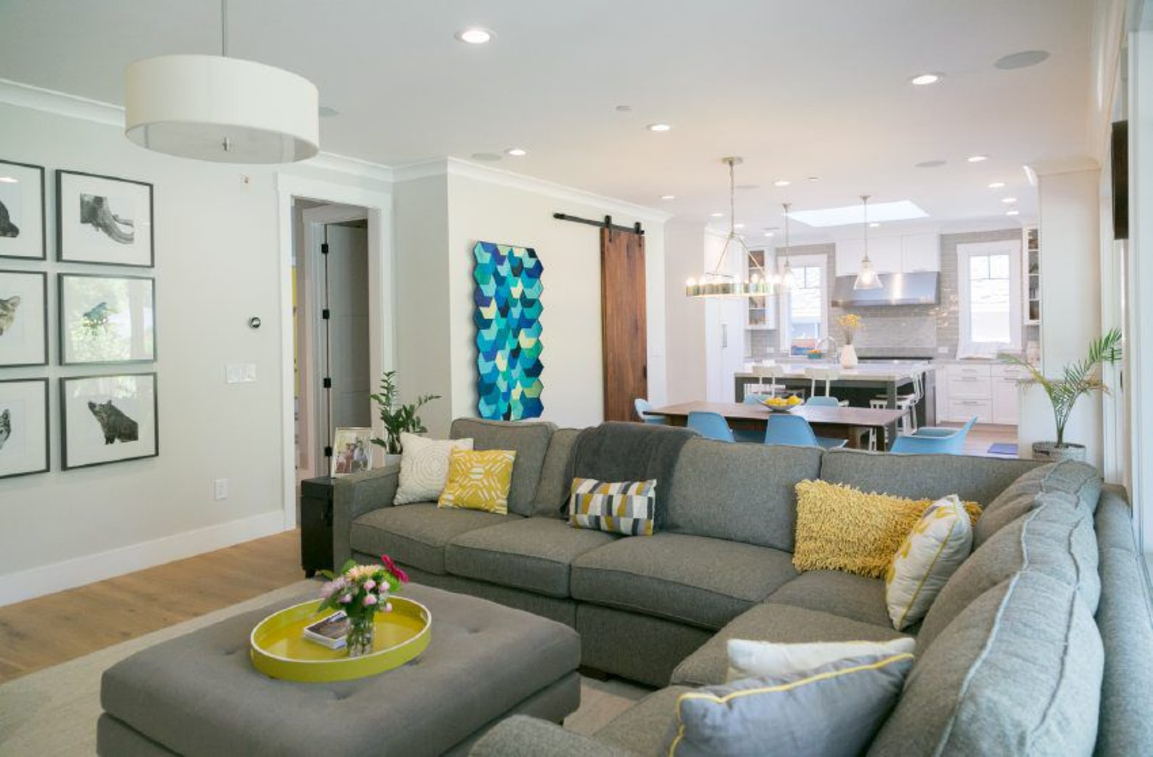A modern living room with a large sectional sofa, a round coffee table, and artwork on the walls, leading into a brightly lit kitchen space.