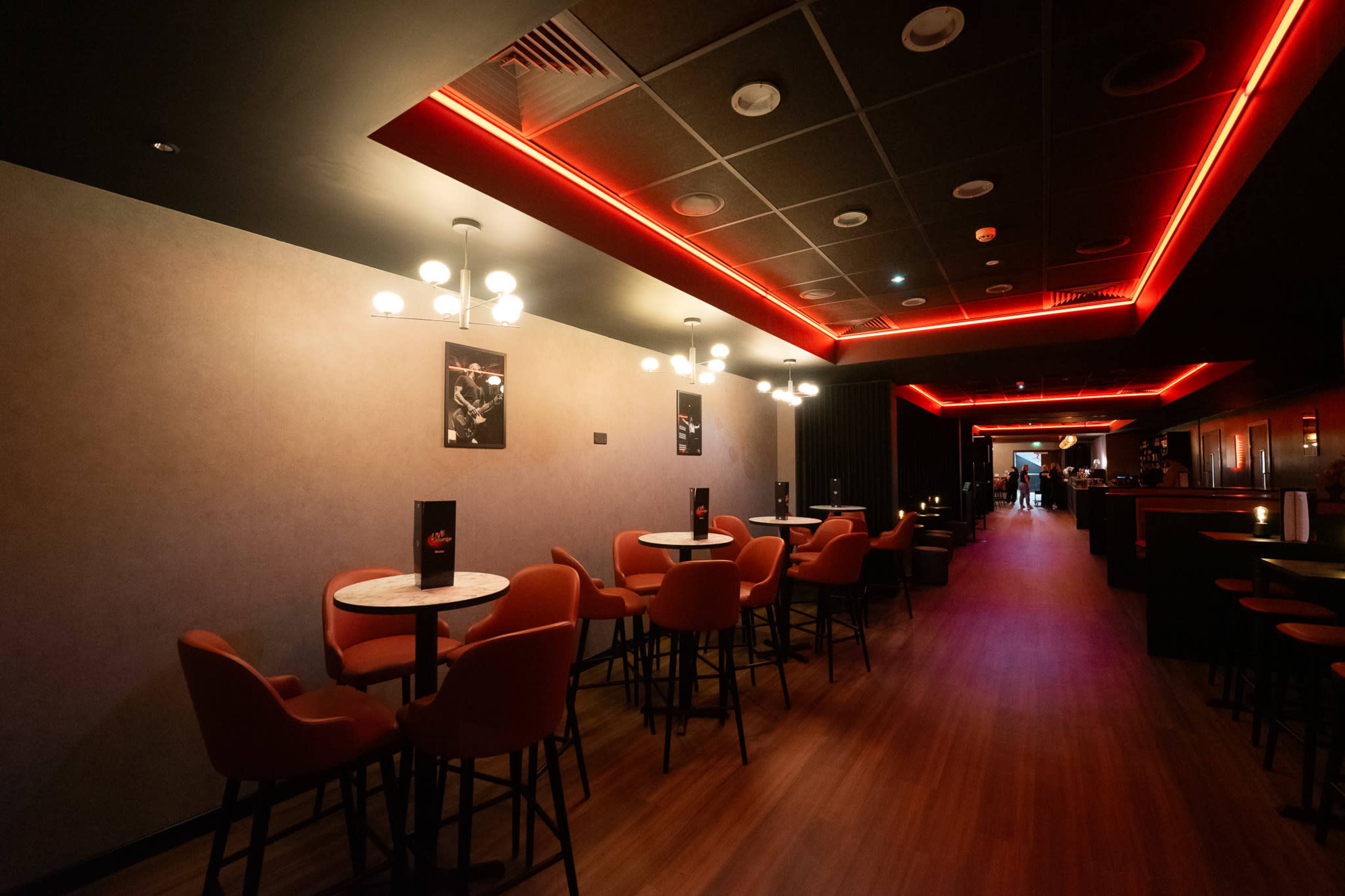 The image shows a modern bar interior with red accent lighting, featuring several round tables surrounded by high chairs along a sleek wooden floor.