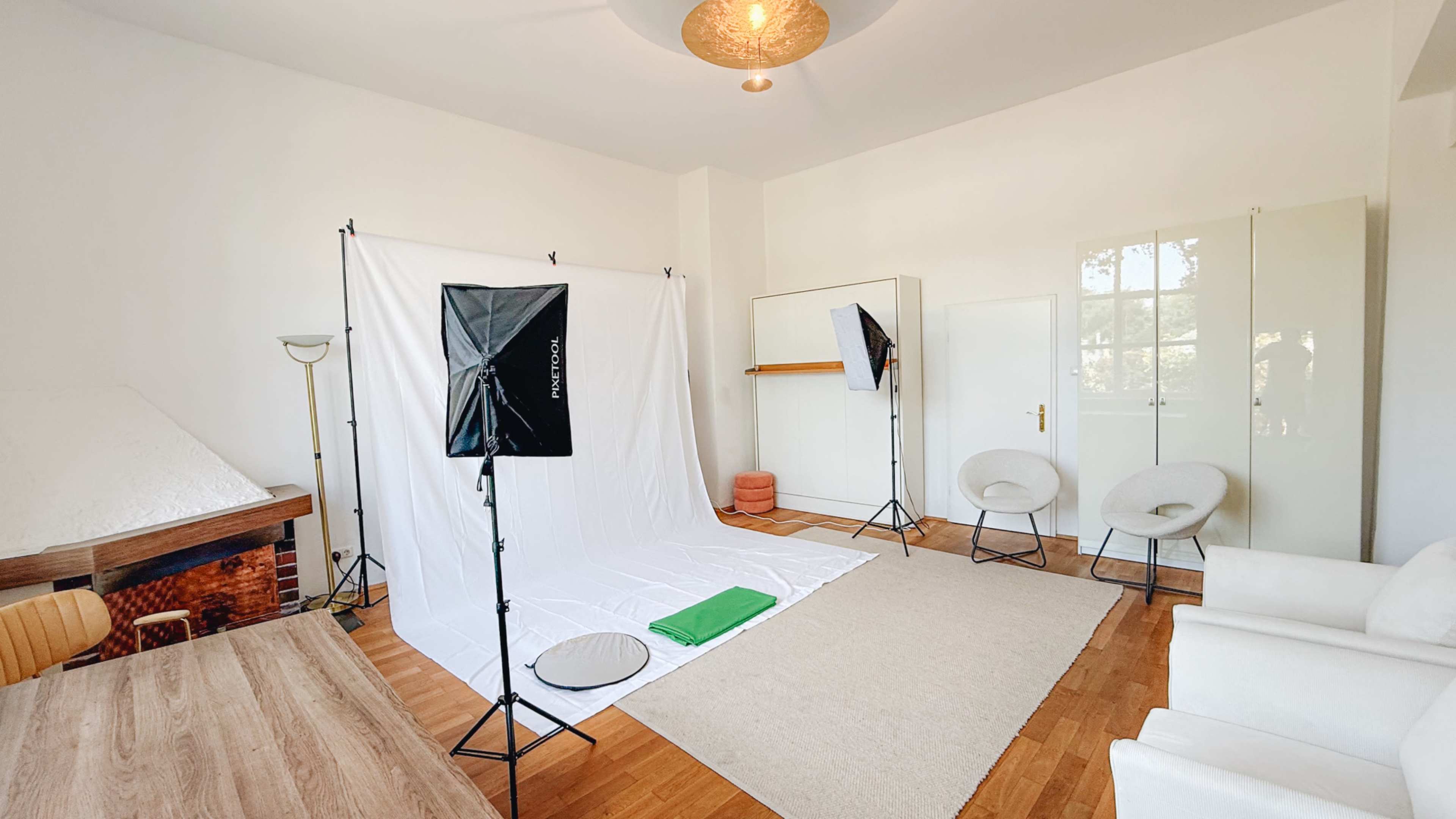 The image shows a bright studio space with white walls, a large white backdrop, two light stands, a rug, a table, and a few chairs arranged neatly.