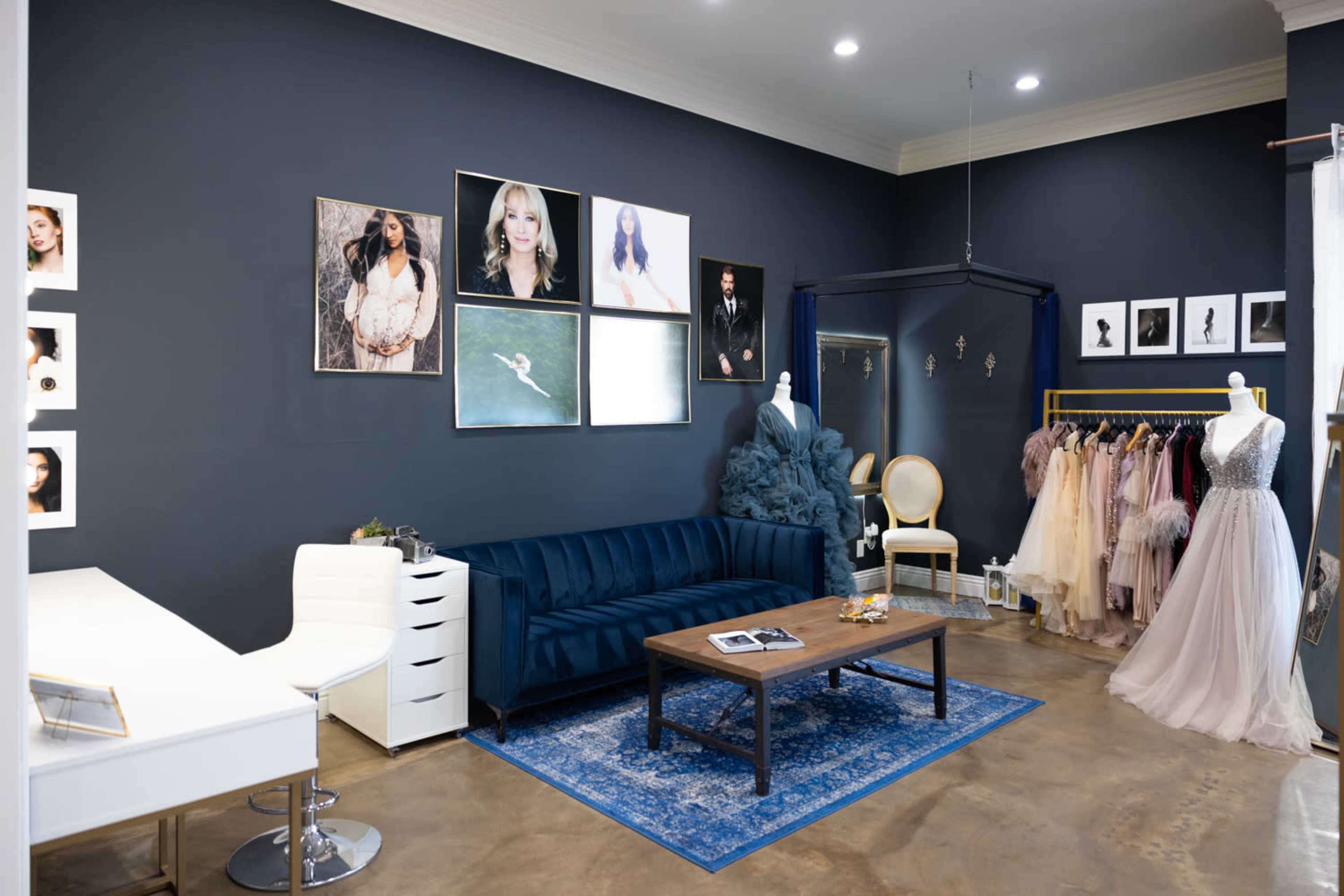 A stylish studio featuring a dark blue wall, a blue velvet sofa, a wooden coffee table, and various framed photographs, along with several elegant dresses displayed on mannequins.