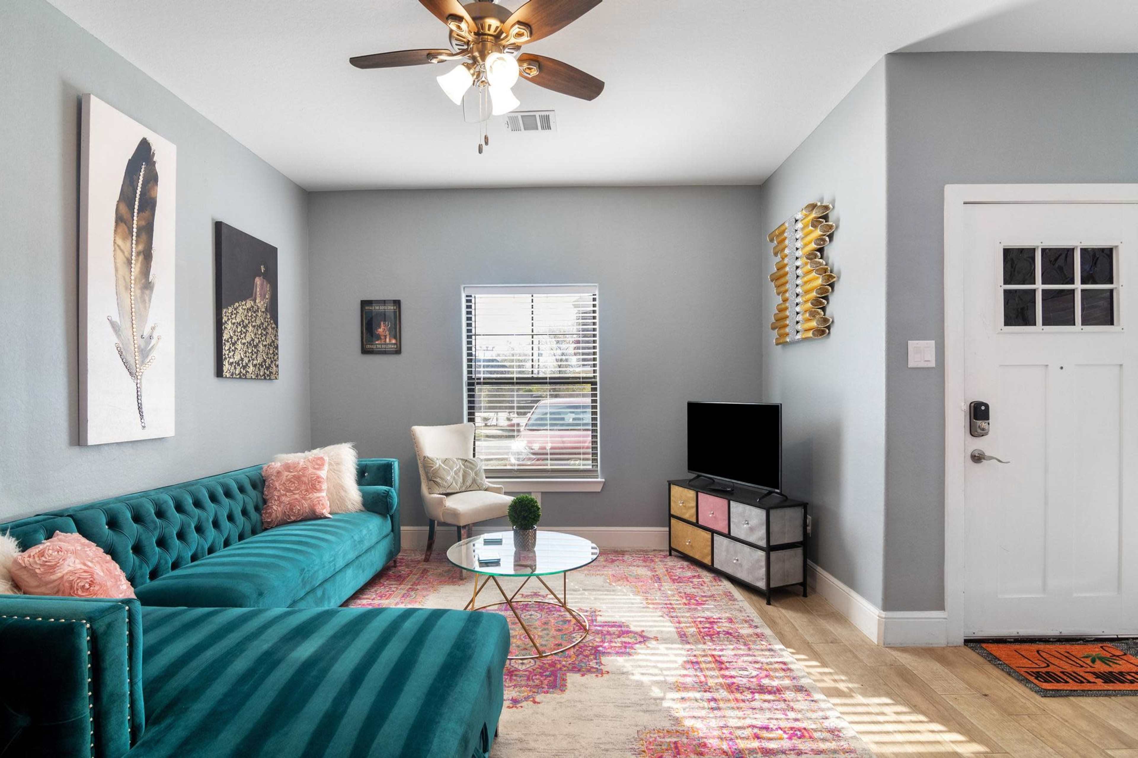 The living room features a teal velvet sofa, a round glass coffee table, and a television on a multi-drawer stand, with artwork on the walls and a window allowing natural light.