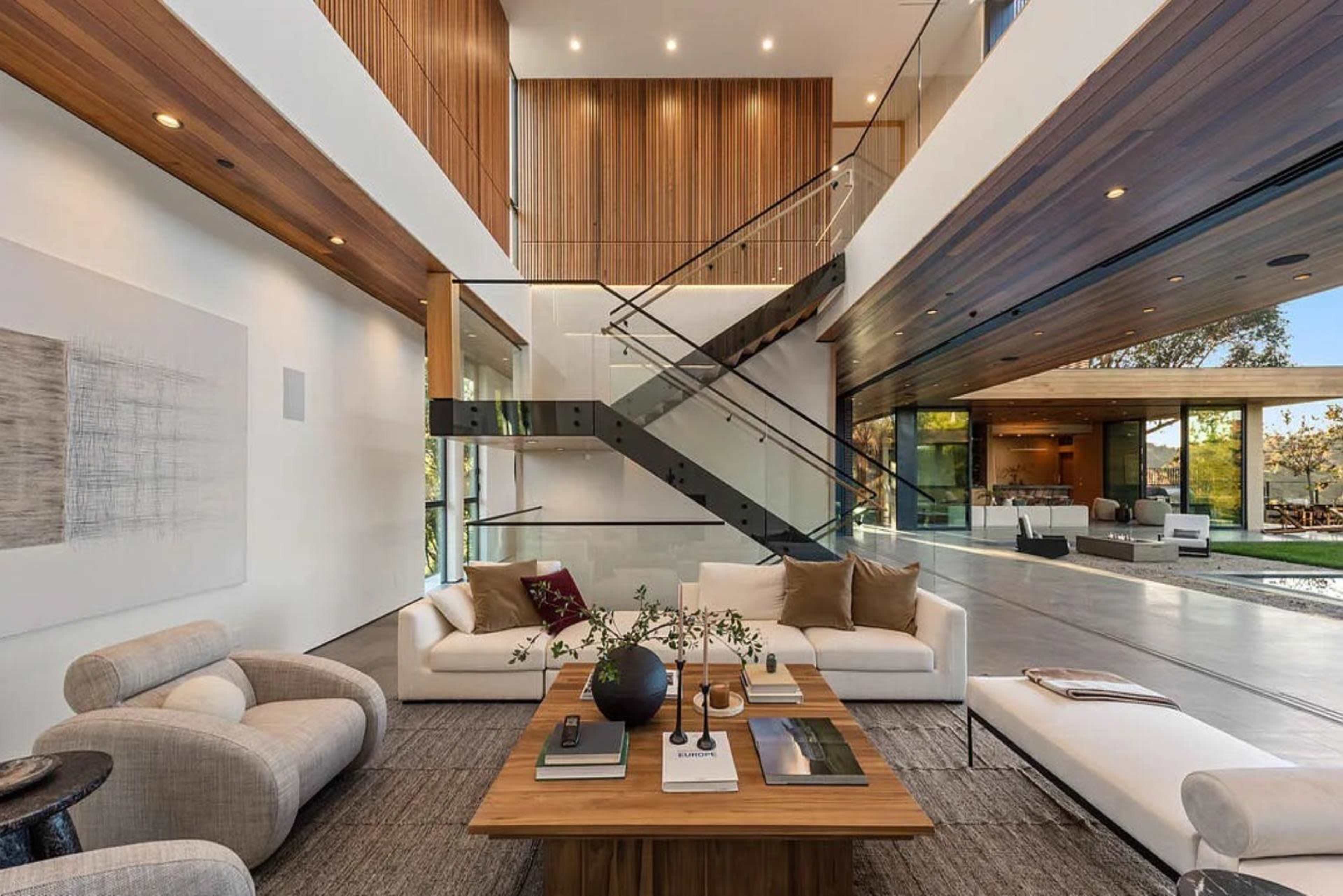 The image depicts a modern living room with a spacious layout, featuring a large coffee table, minimalist furniture, and a staircase with a glass railing.