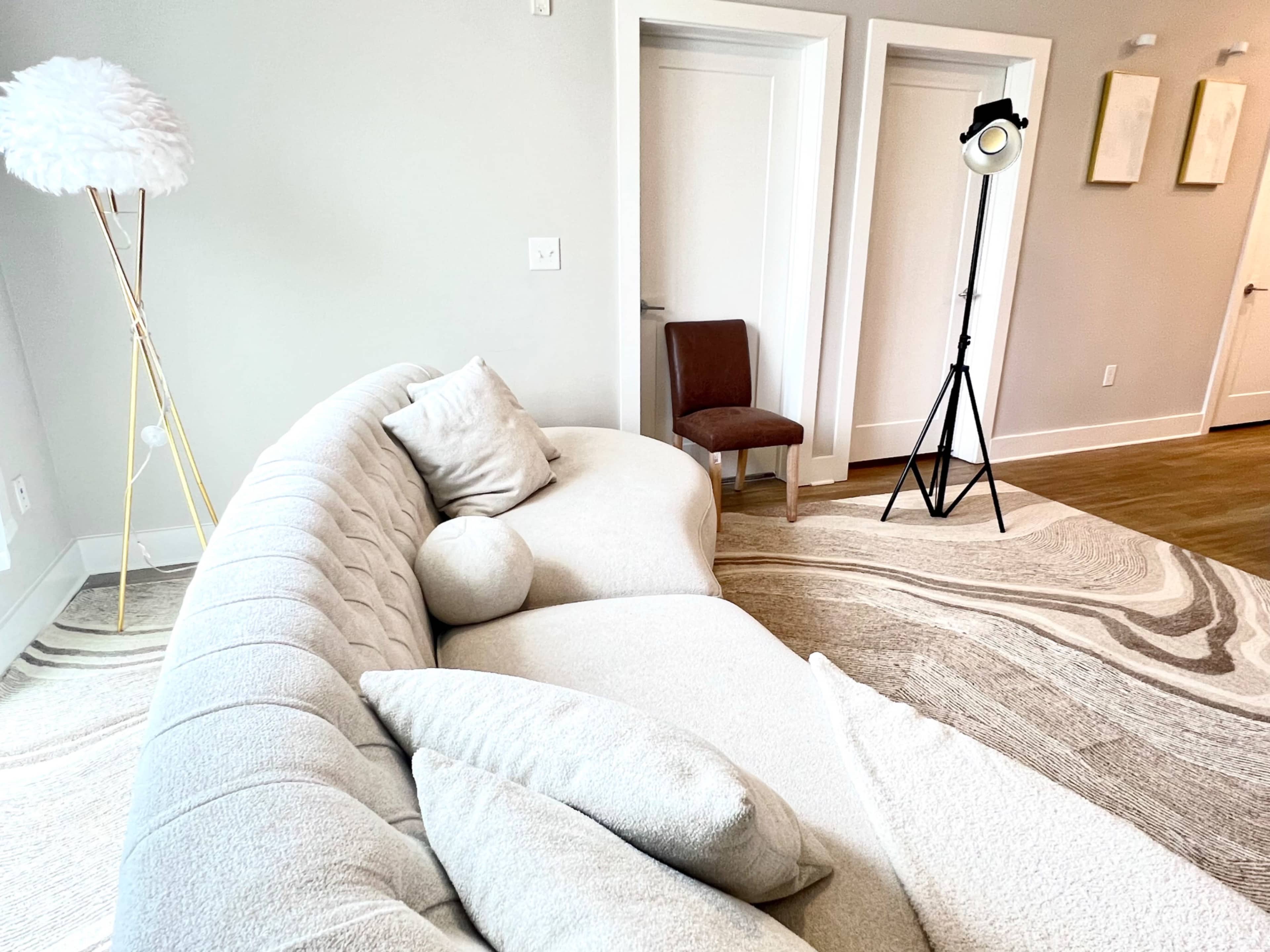 A curved, light-colored sofa is positioned next to a wooden chair and a floor lamp in a minimalist living room.