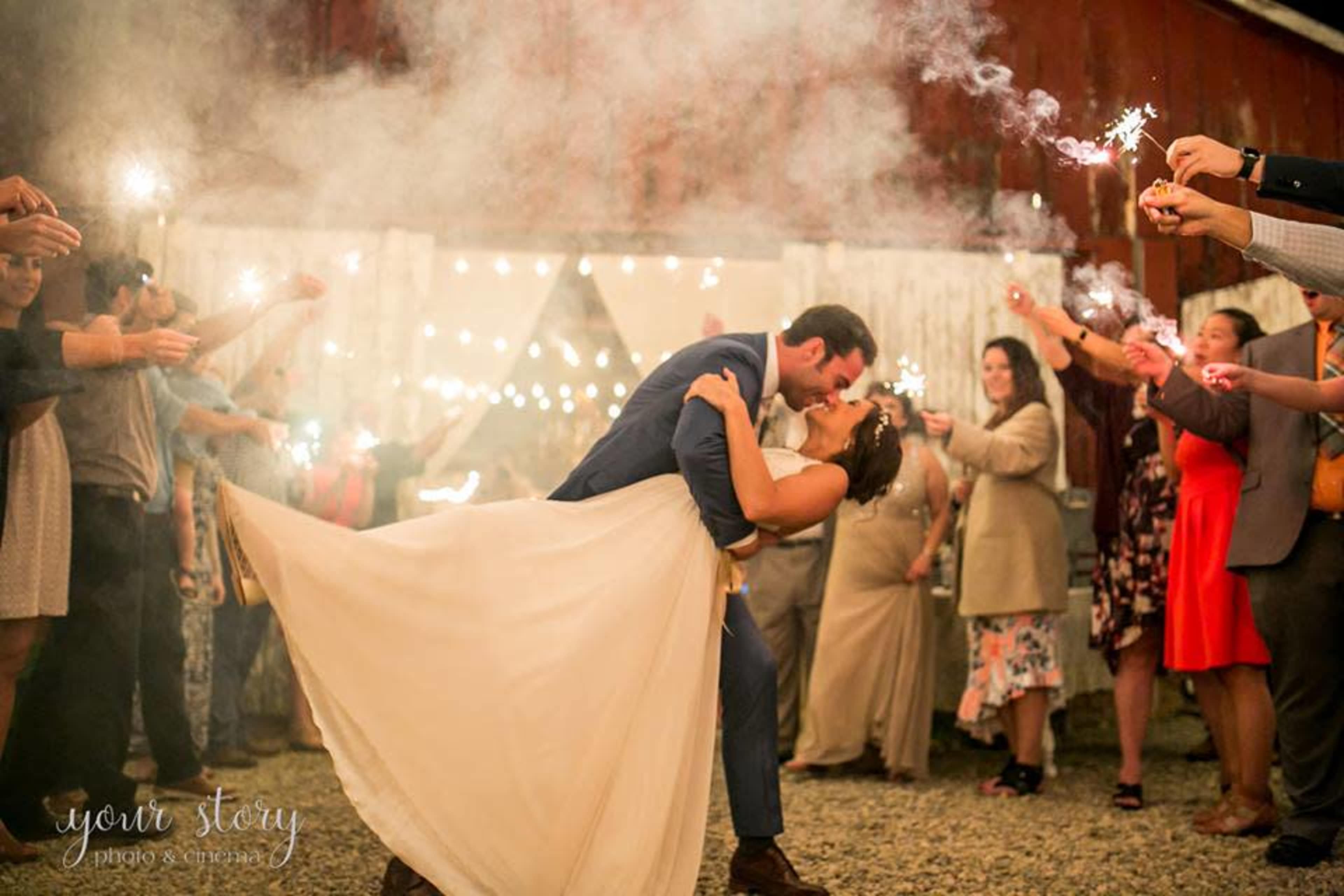 A bride and groom share a kiss while surrounded by wedding guests holding sparklers in front of a rustic barn.