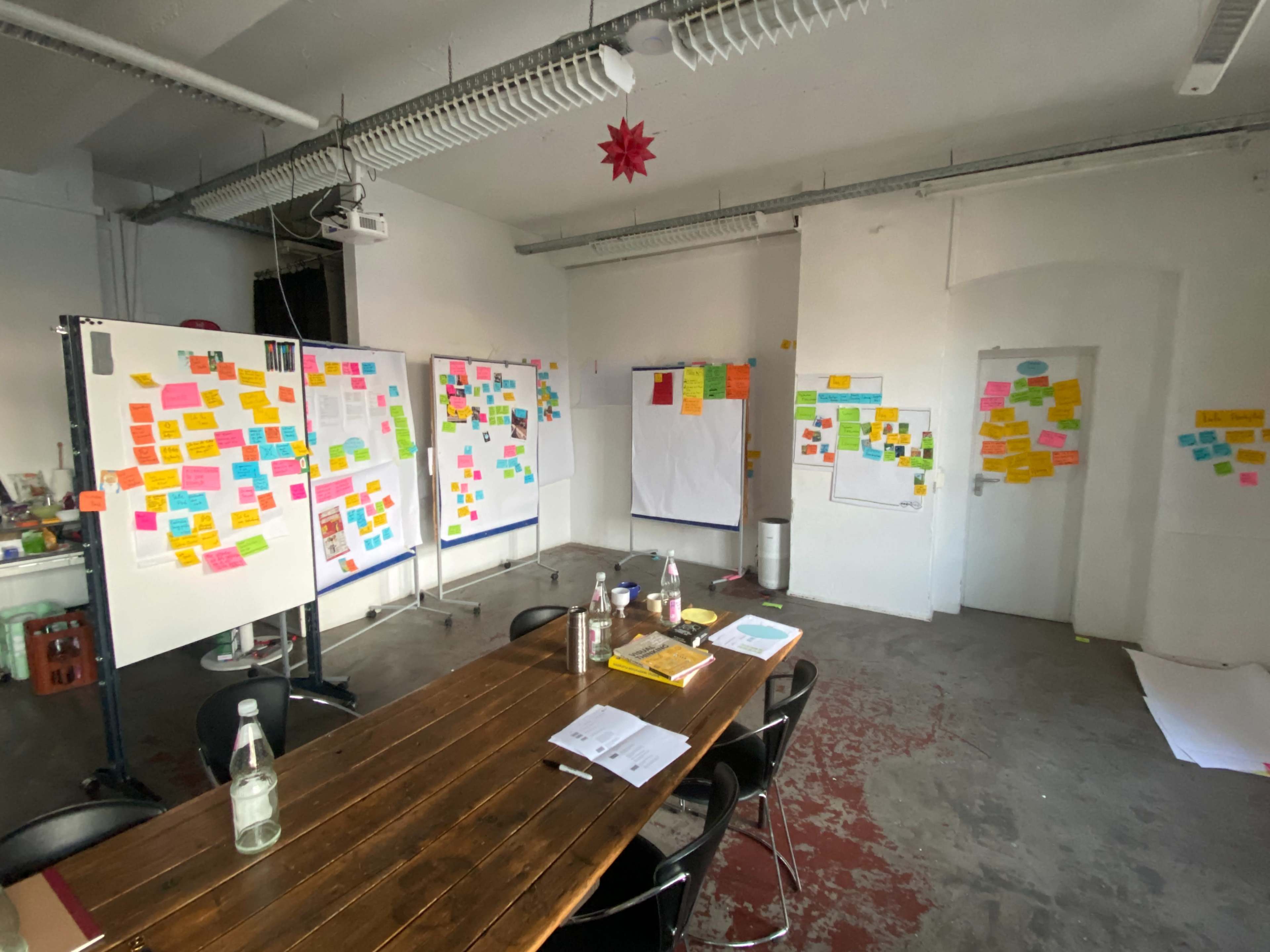 A meeting room features a long table surrounded by chairs, with walls covered in colorful sticky notes and a large blank sheet of paper on display.
