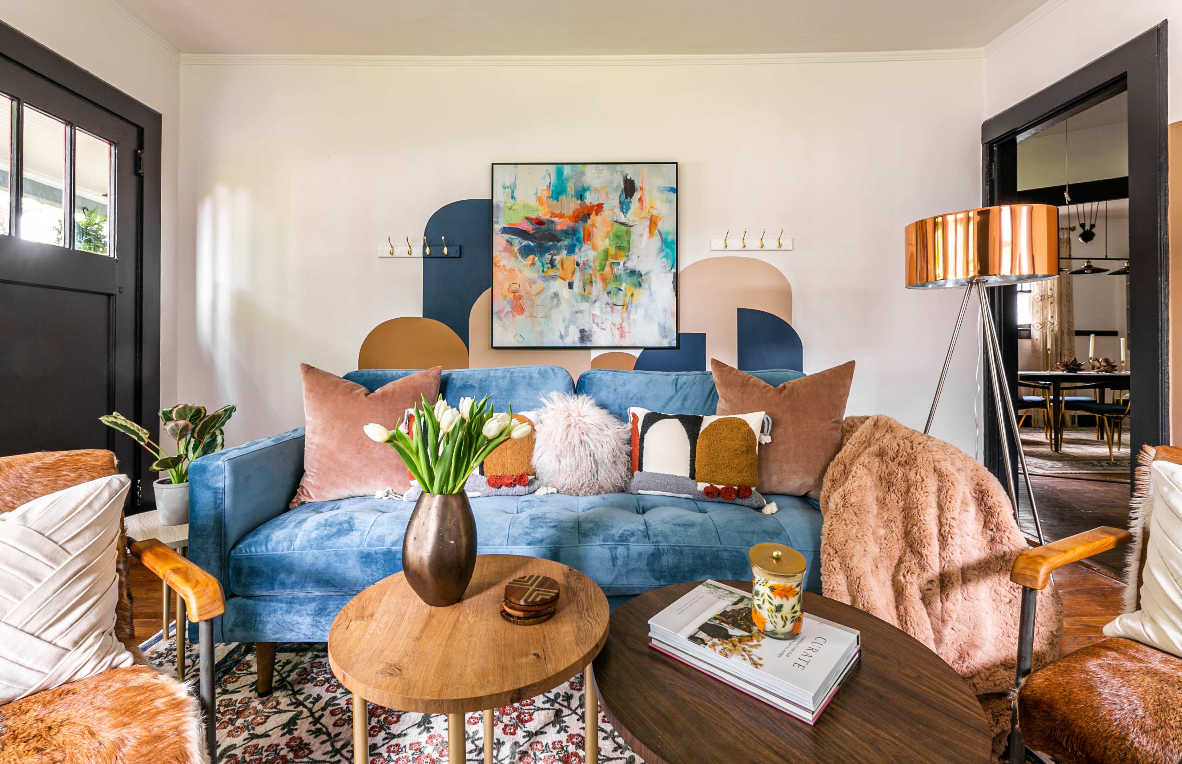 A cozy living room with a blue couch adorned with colorful pillows, a round wooden coffee table, and a lamp, all set against a backdrop of abstract art on the wall.